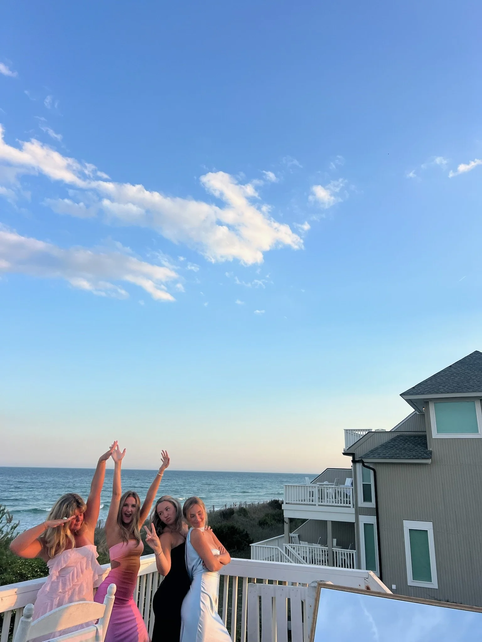 A group of five young women standing on a white balcony near the beach, making playful poses with hands raised towards the sky, with the ocean, blue sky, and a house visible in the background.