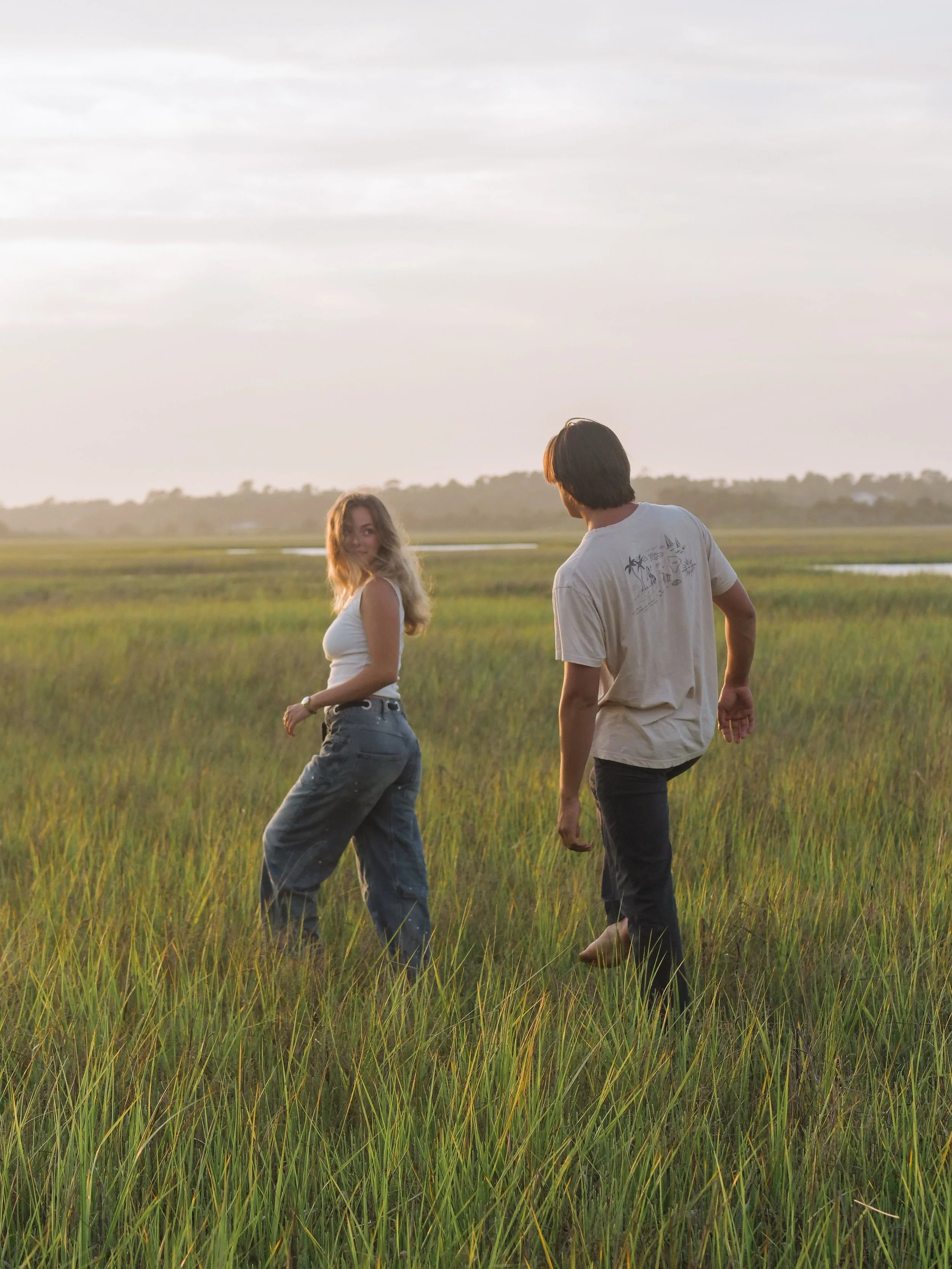A young woman with wavy blonde hair and a white shirt smiling and looking over her shoulder while walking in a grassy field at sunset, with a young man with dark hair and a light-colored shirt walking nearby.