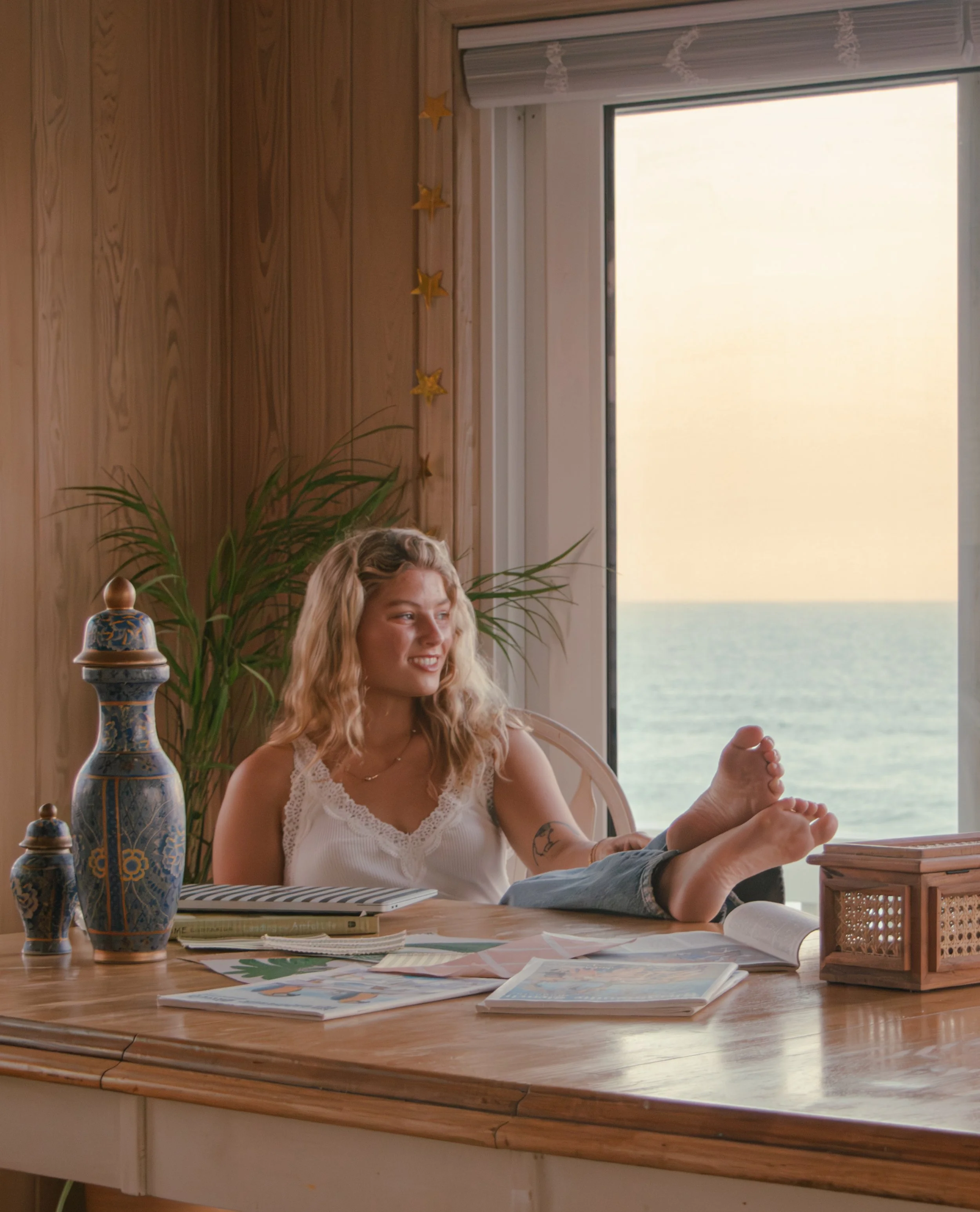 A woman with long blonde hair smiling while sitting at a wooden table. The table has multiple coloring books, papers, and a wooden box. There are decorative jars and little yellow star ornaments hanging on the wall behind her. A large window shows a view of water and sky.