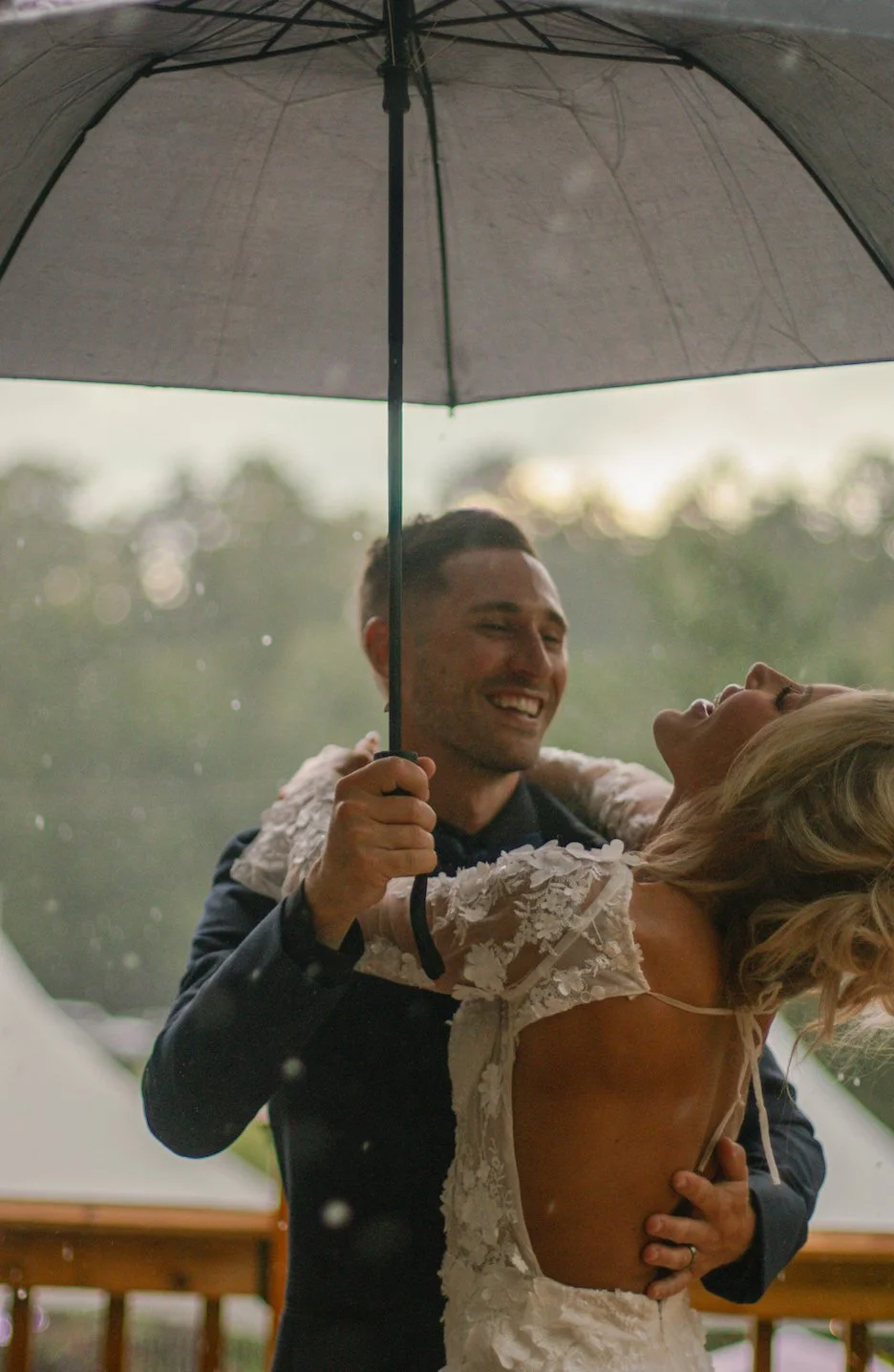 A couple dancing under a black umbrella outdoors, with blurred trees in the background. The man is holding the umbrella while the woman, wearing a white lace dress, leans back happily.