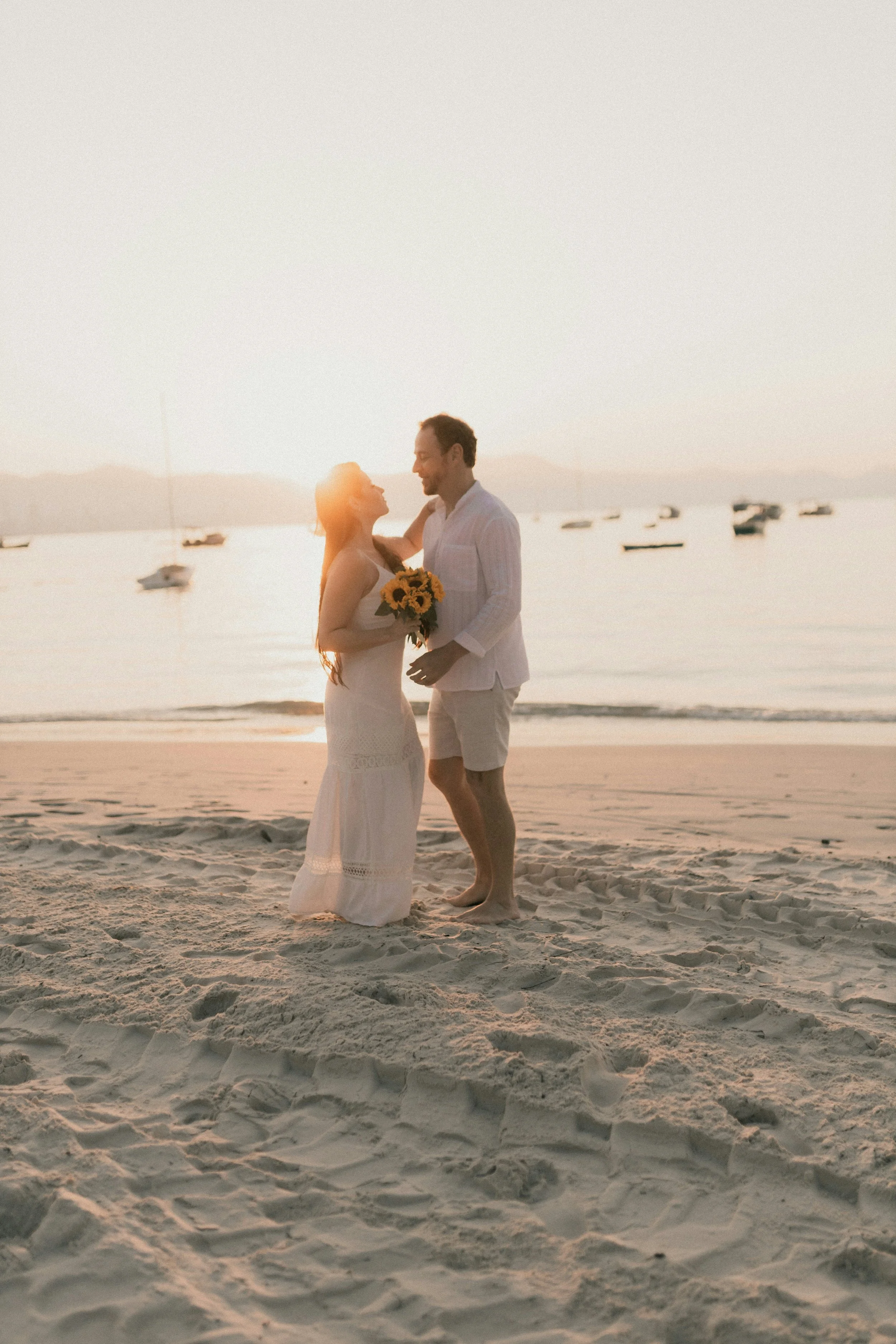 A wedding couple is standing on a sandy beach during sunset, gazing at each other, with the woman holding a bouquet of sunflowers, and boats anchored on the water in the background.