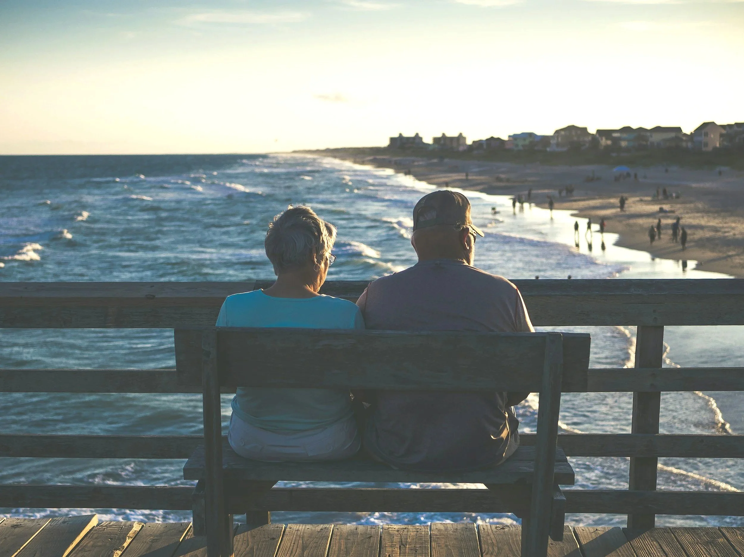 Two elderly people sitting on a wooden bench on a pier, watching the ocean at sunset. Houses are visible in the distance along the coast.