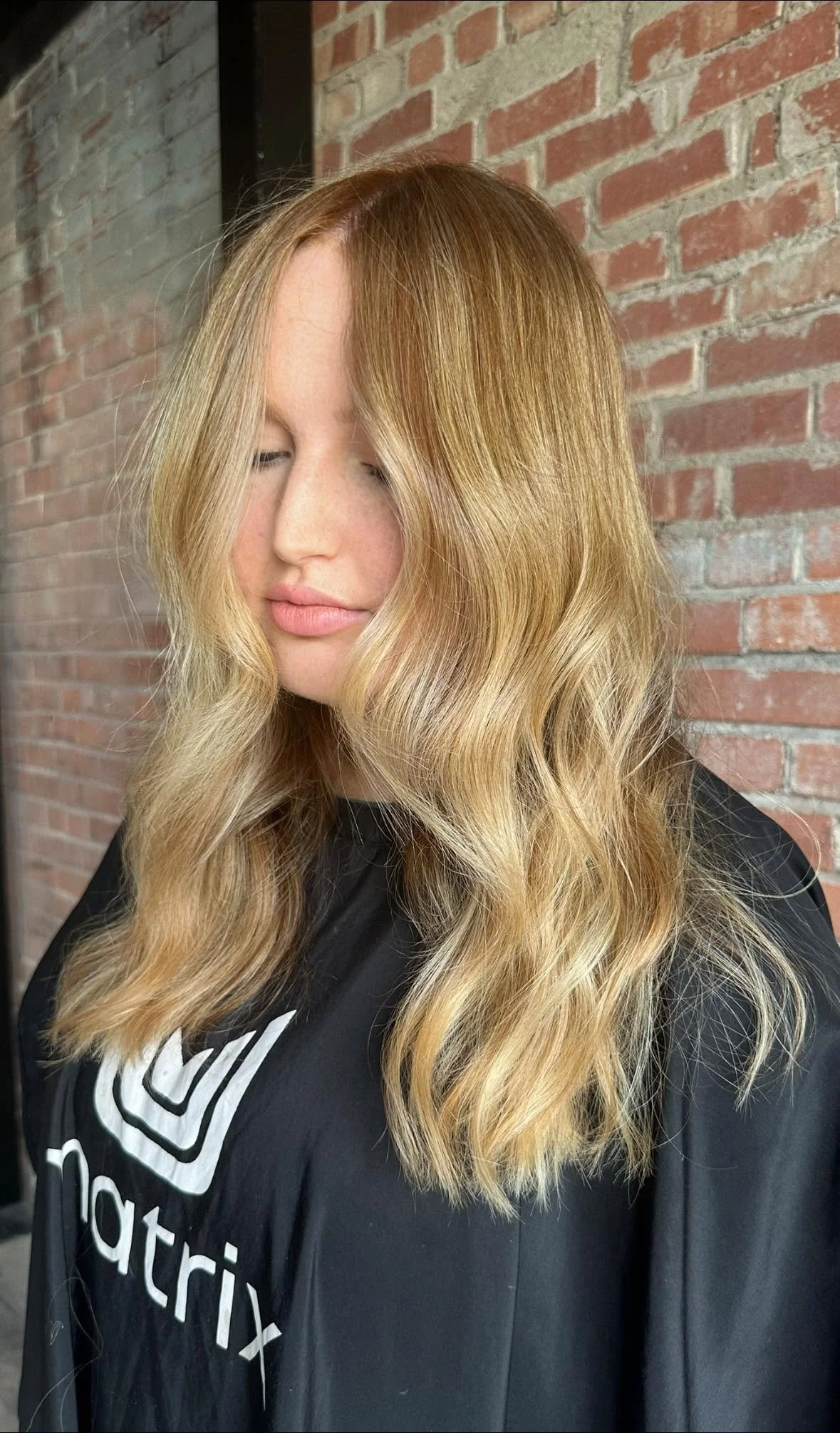 A young woman with long, wavy, blonde hair with darker roots and a side part, standing in front of a brick wall. She has fair skin, closed eyes, and light pink lipstick, wearing a dark salon cape.