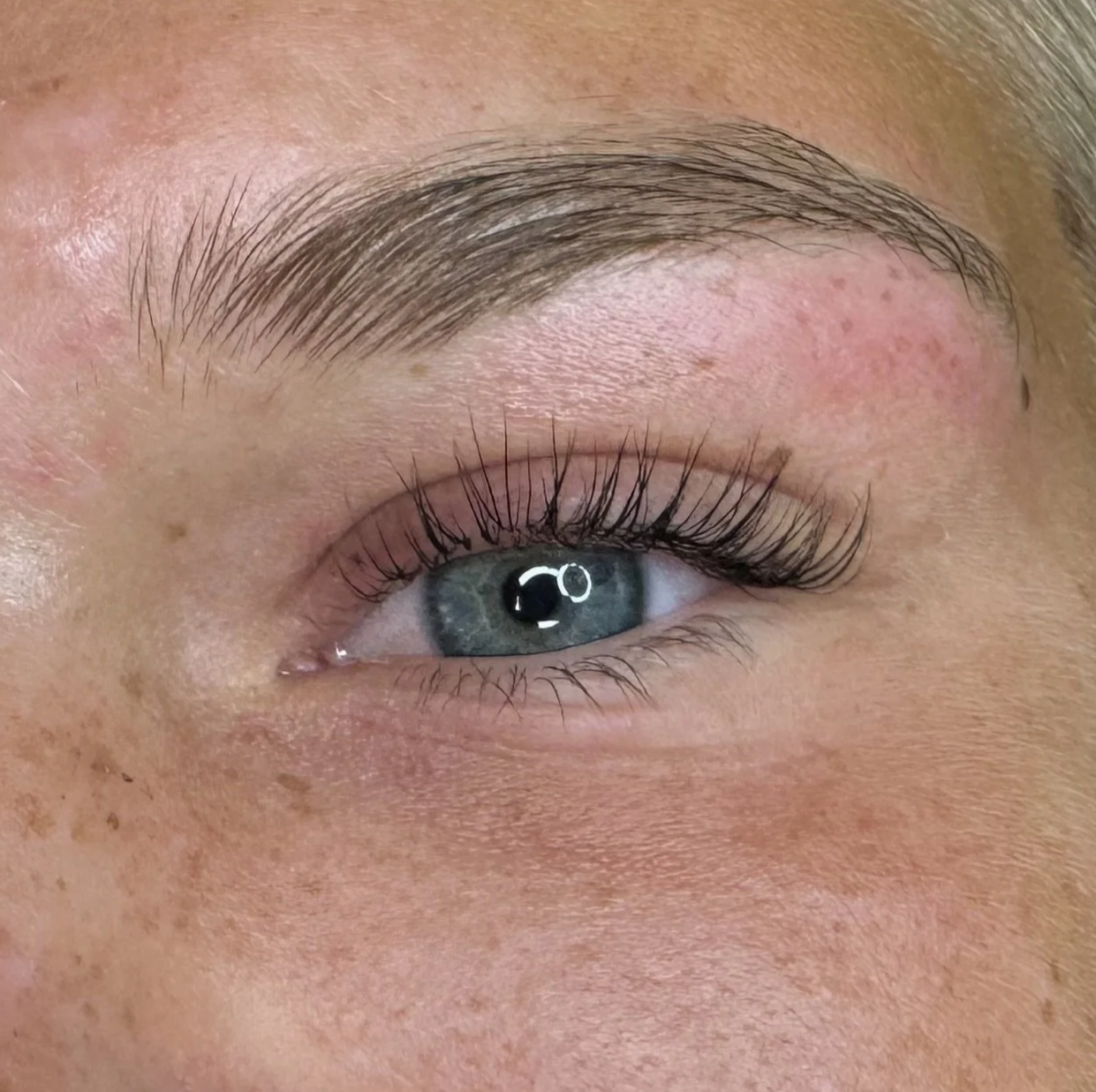 Close-up of a person's eye with well-groomed eyebrow, long eyelashes, and blue-gray iris, reflecting light.