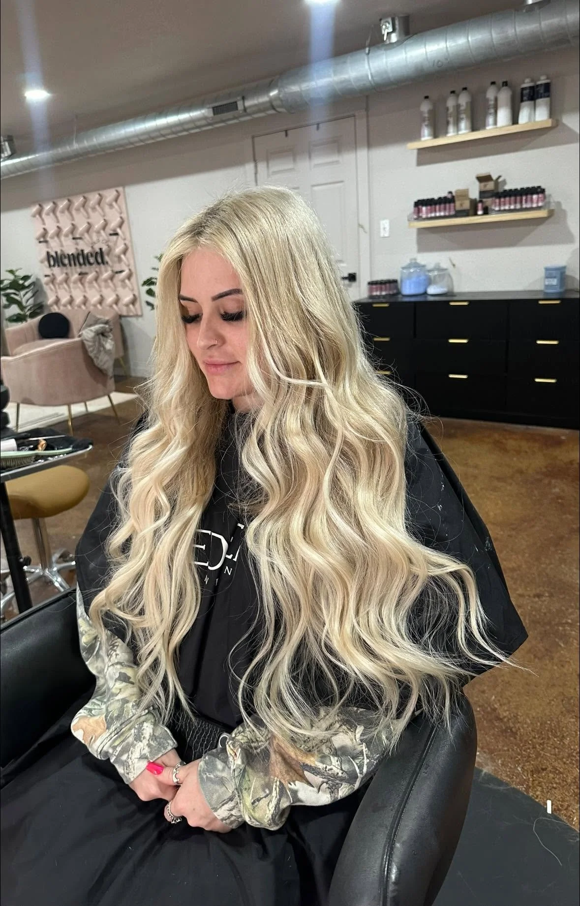 A woman with long, wavy blonde hair sitting in a salon chair, looking down with her eyes closed. The salon has a modern interior with black cabinets, shelves with hair products, and a pink decorative wall with the word 'blended.'