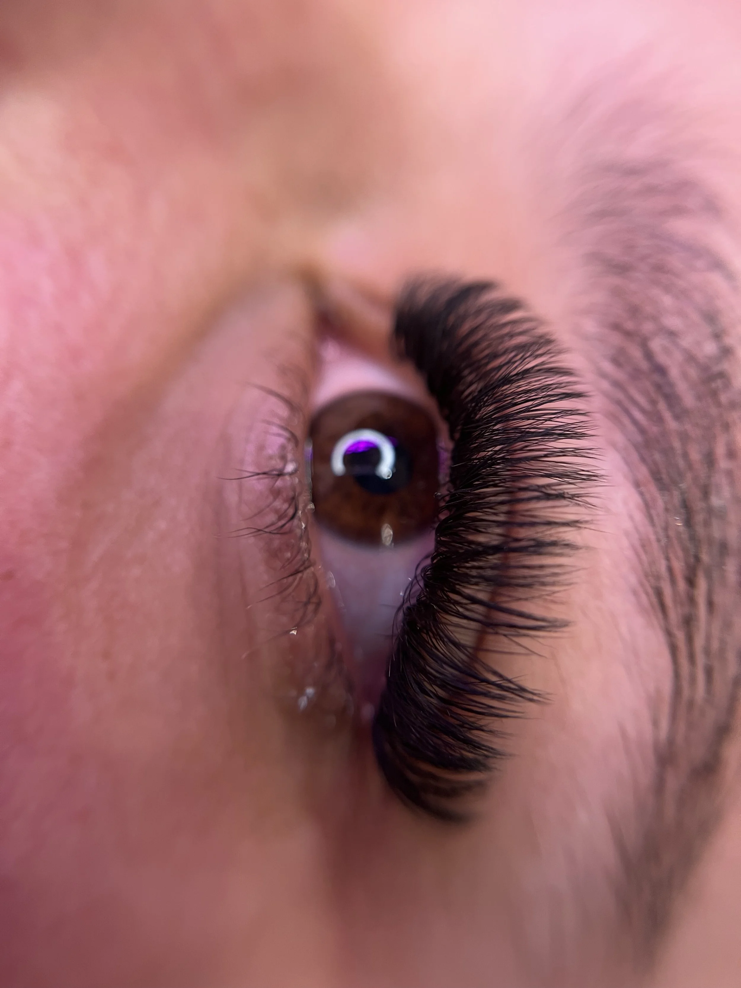 A close-up photo of a person's eye showing long, dark eyelashes and a reflection of a circular light in the eye.