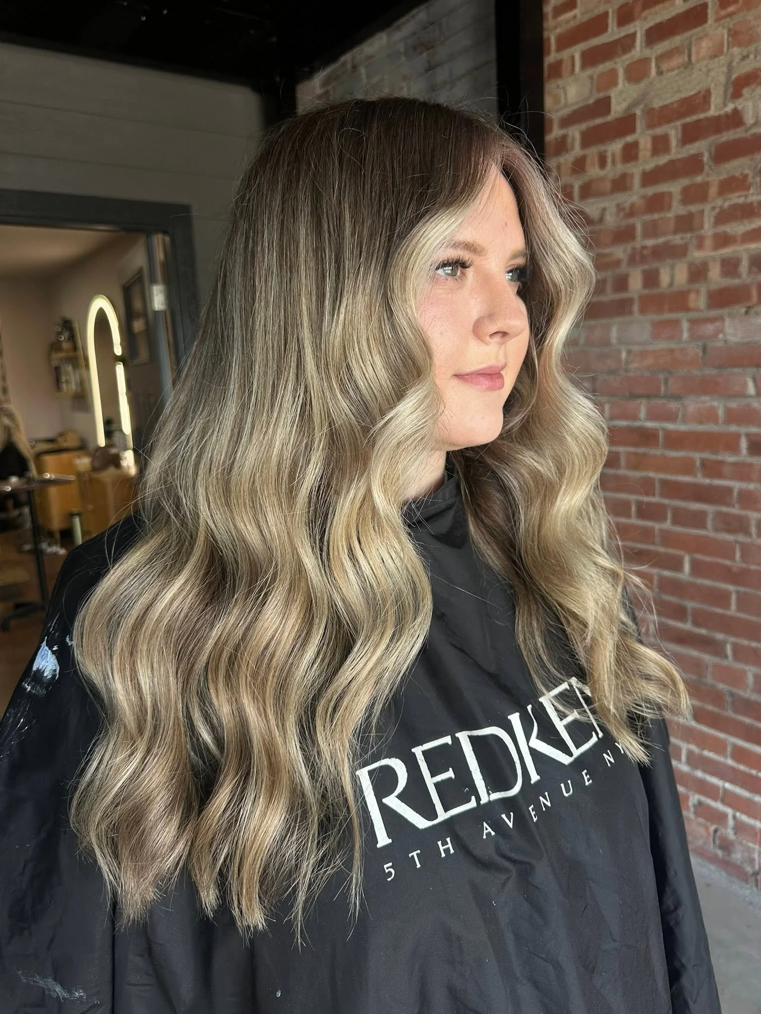 A woman with long, wavy blonde hair with darker roots, wearing a black cape with white lettering, sitting in front of a brick wall in a salon.
