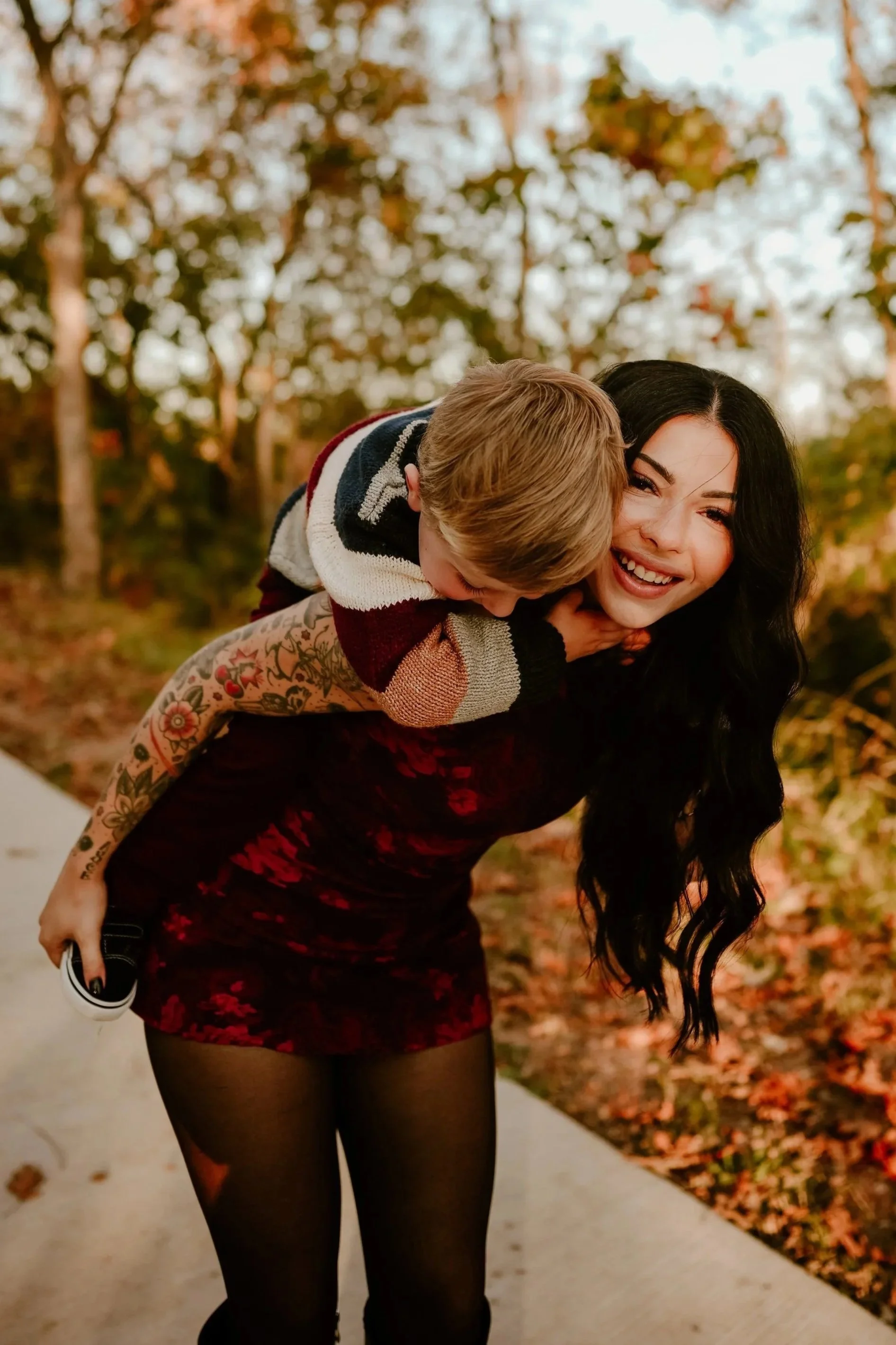 A woman with long black hair and tattoos on her arm, wearing a red and black floral dress with black tights, giving a piggyback ride to a young boy with blonde hair, outdoors with fall foliage in the background.