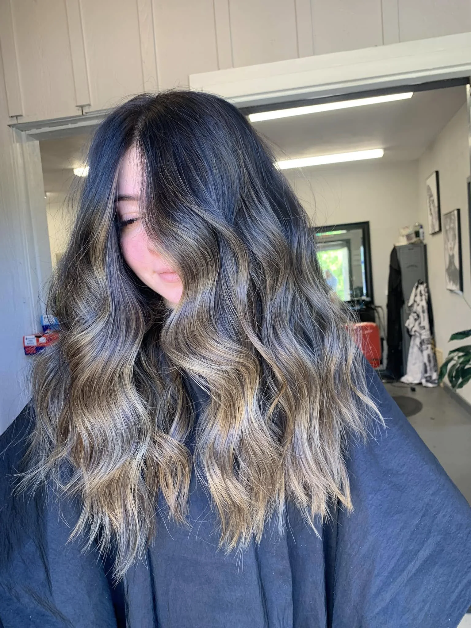 Person with long, wavy hair that is dark at the top and gradually becomes blonde towards the ends, indoors with a background of white paneled walls, fluorescent lighting, and framed pictures.