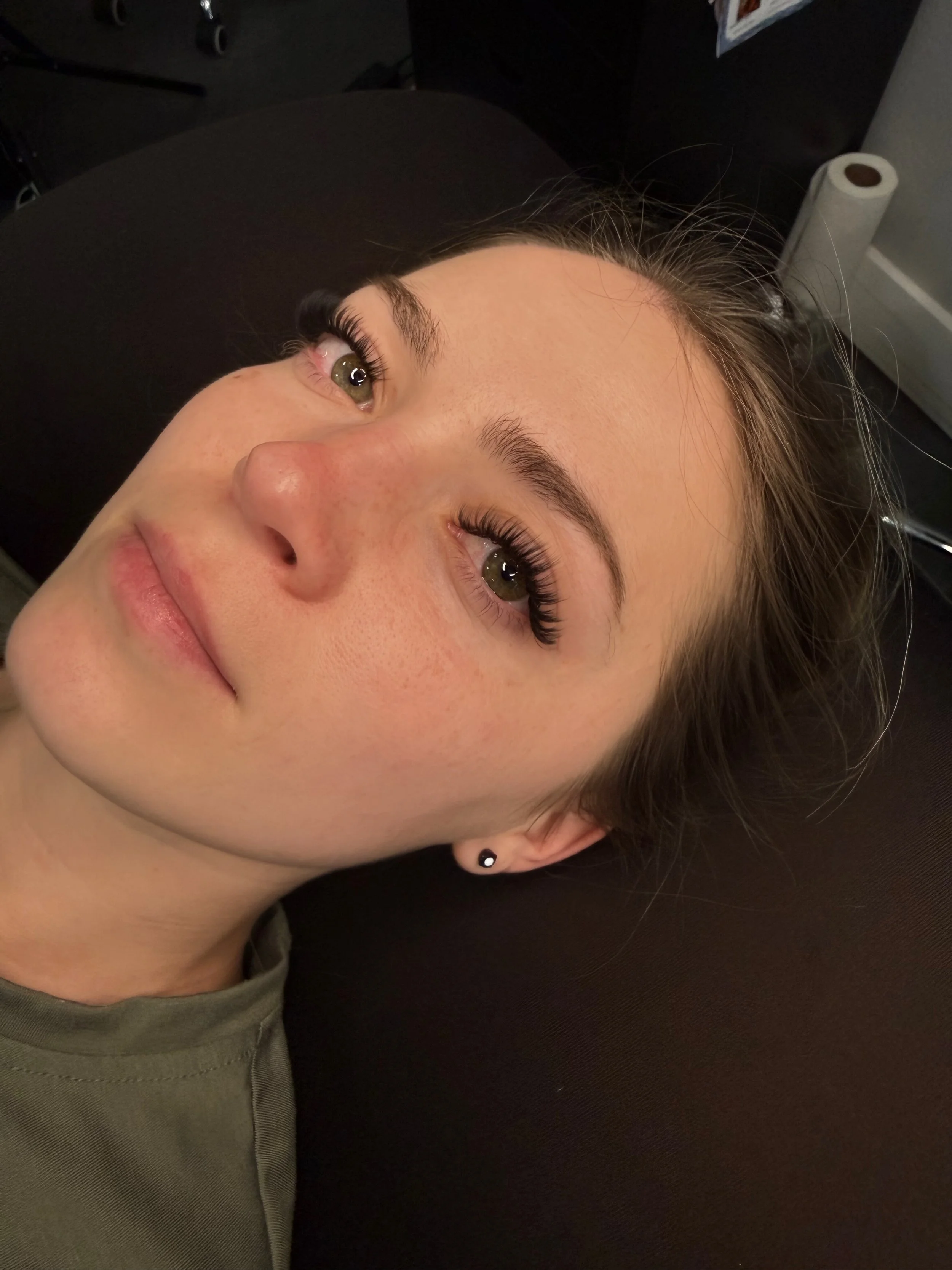 Close-up of a woman lying down with dark hair, makeup, and earrings, looking upwards, in an indoor setting.