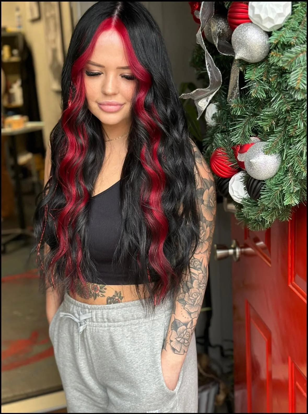 A woman with long black hair with red highlights, tattoos on her left arm and stomach, wearing a black crop top and gray sweatpants, standing next to a decorated Christmas tree with red, silver, and white ornaments.