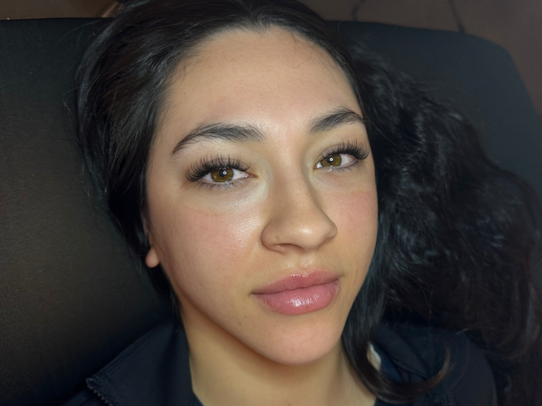 A woman with light brown eyes and dark hair lying on a black surface, looking into the camera, with makeup and well-groomed eyebrows.