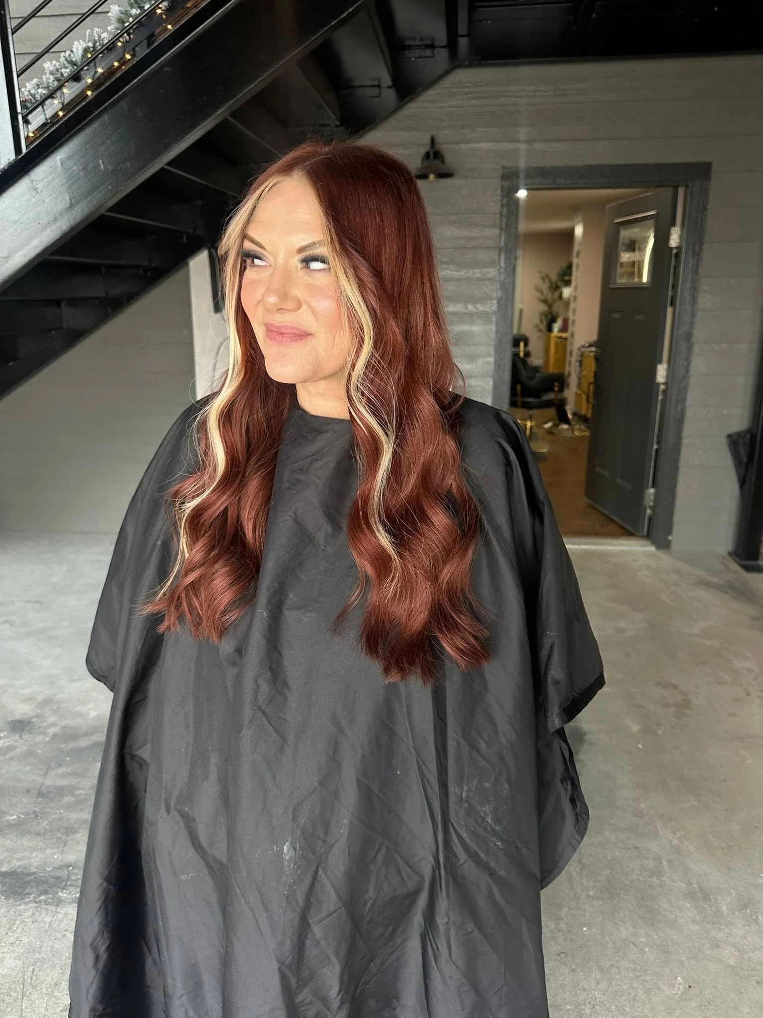 A woman with long, wavy red hair and blonde streaks, wearing a black salon cape, standing in a hair salon.