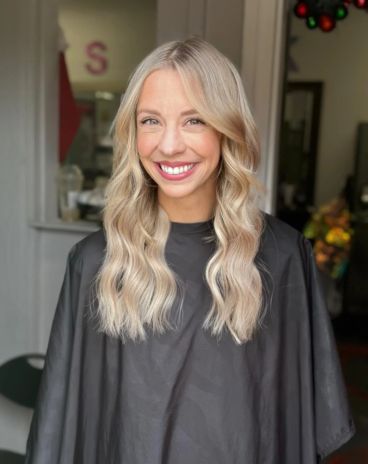 A woman with long, blonde, wavy hair smiling, wearing a black salon cape, standing indoors.