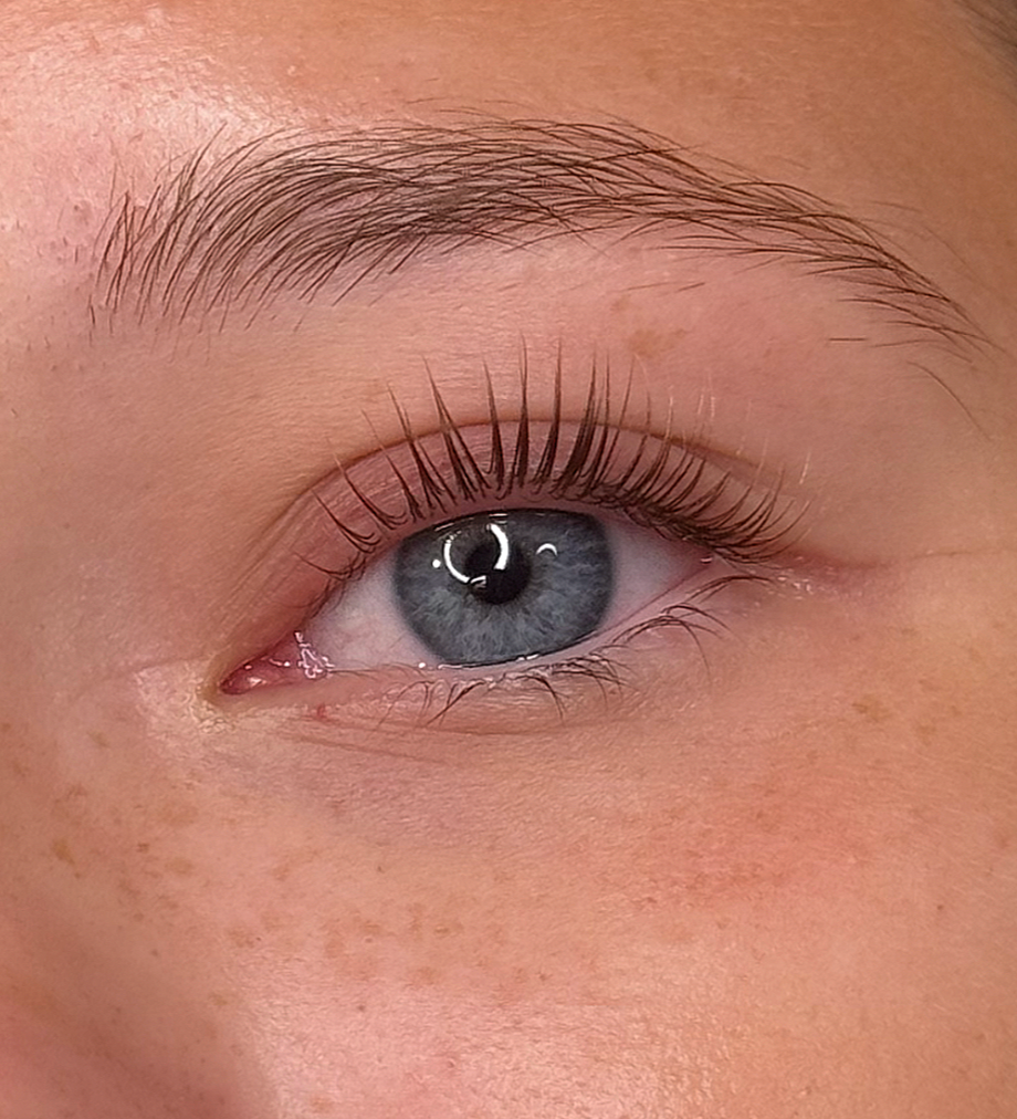 Close-up of a person's blue eye with long eyelashes and light skin, showing a reflection of a ring light in the eye.