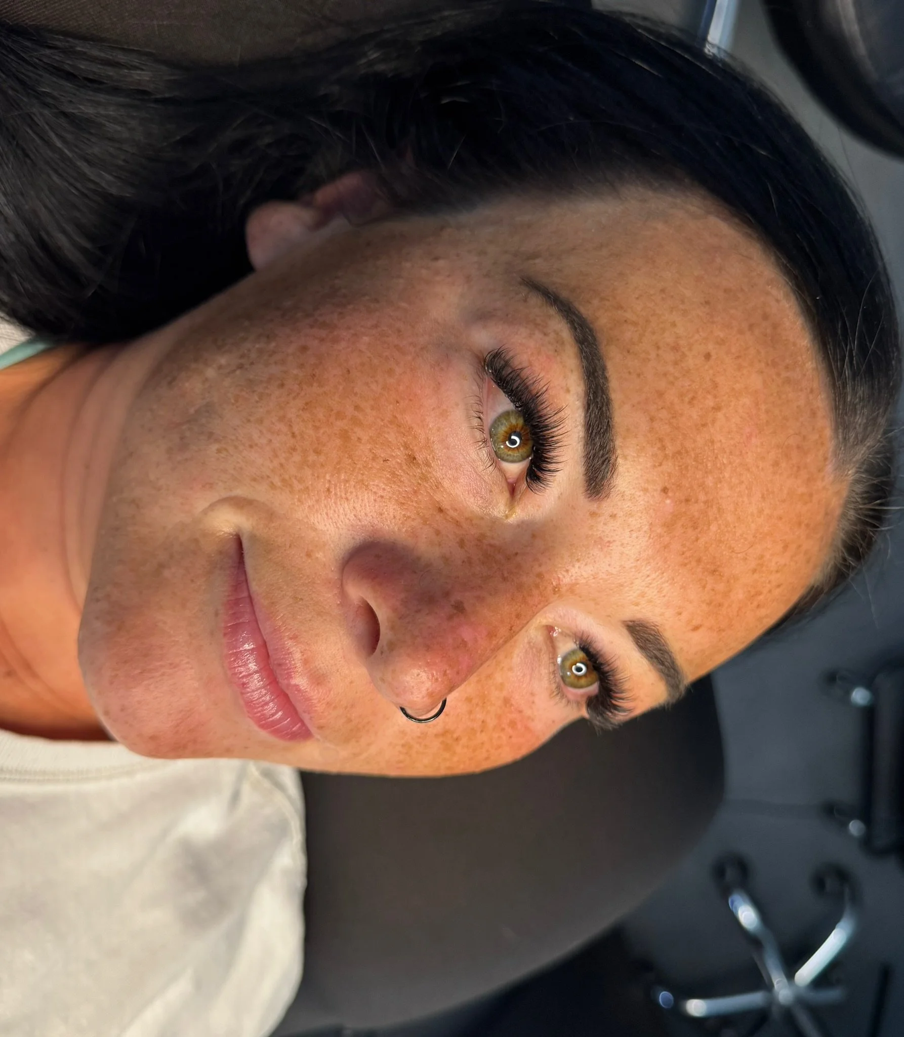 Close-up of a woman with dark hair, freckles, and green eyes, lying down with her face turned to the side, wearing a hoop earring.