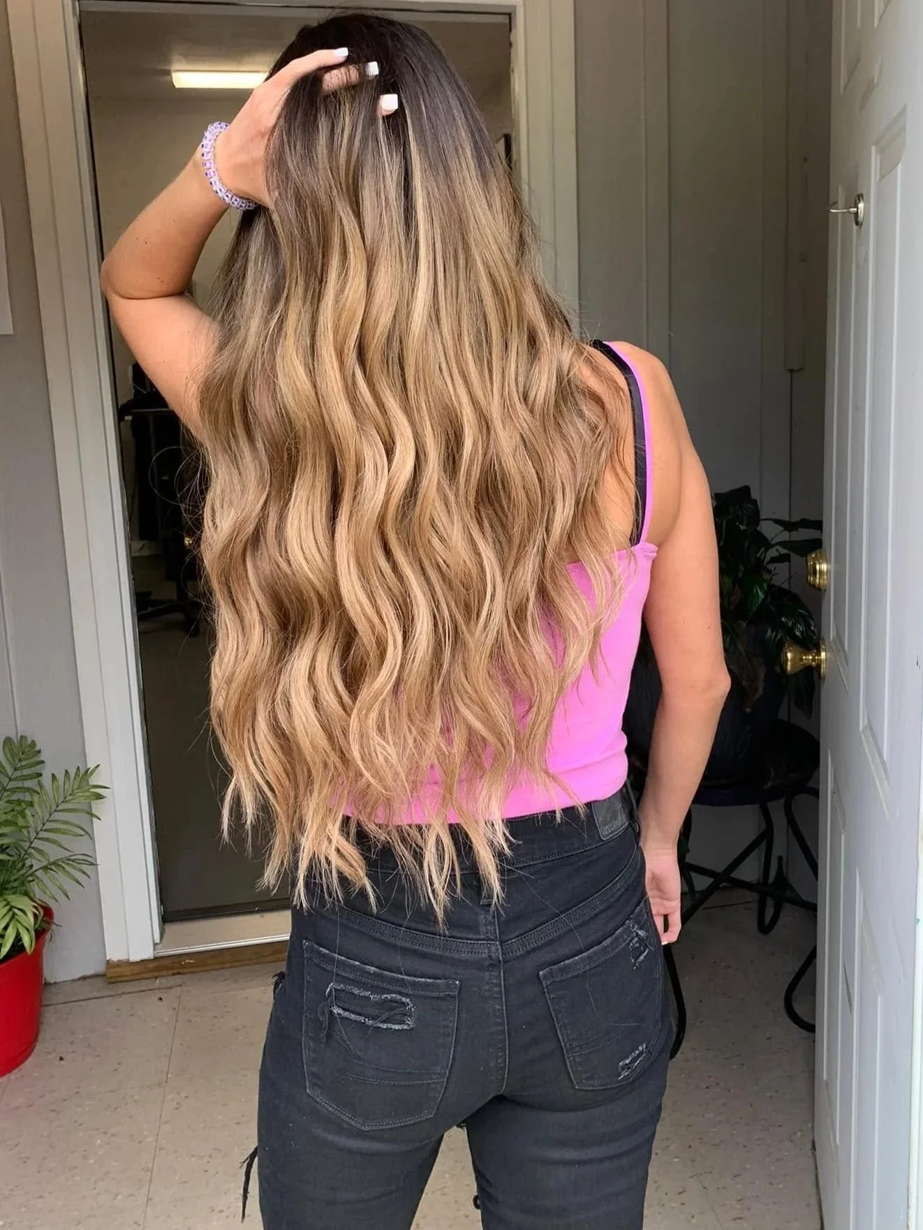 Back view of a woman with long, wavy blonde hair, wearing a pink tank top and black ripped jeans, standing at the doorway of a house, with her left hand resting on her head.