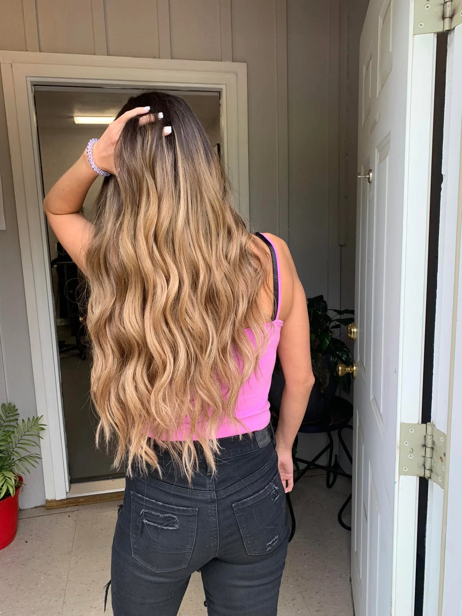 A person with long wavy hair, light brown at the top and blonde at the ends, wearing a pink tank top and black jeans, is standing at a doorway with their back to the camera. Their right hand is touching their head, and they are inside a room that app