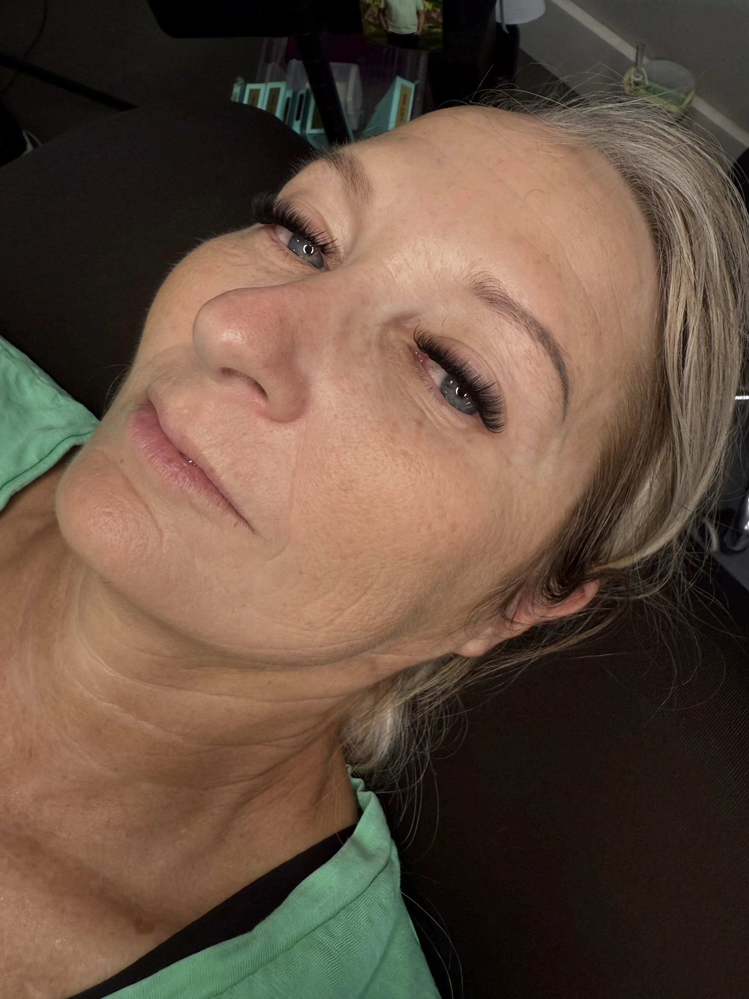 A close-up of a woman lying down, showing her face with light-colored hair, blue eyes, and freckles, wearing a green top. The background includes some objects and people.