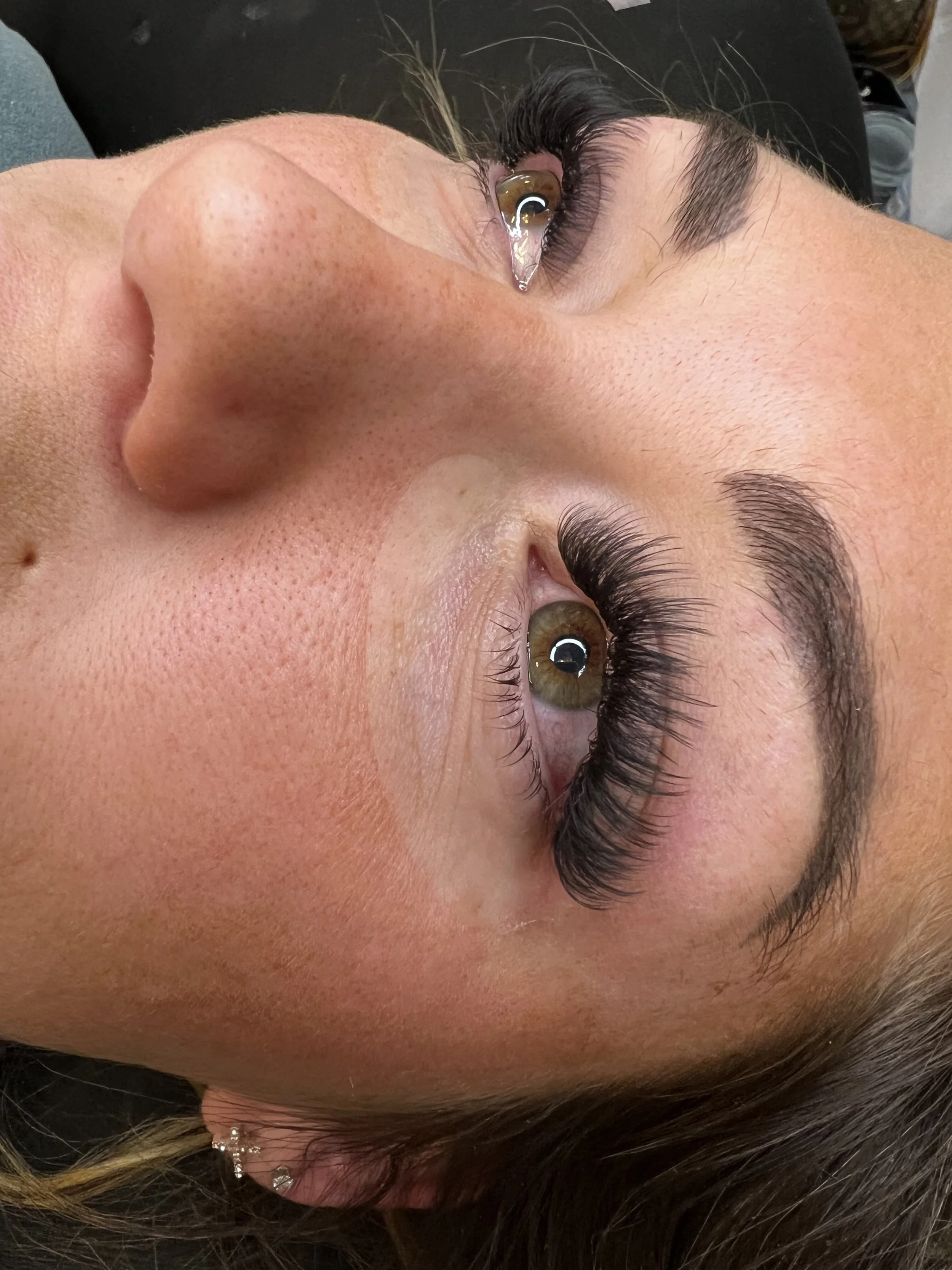 Close-up of a woman's face lying on her side, showing her eye with long eyelashes, and her nose. Her skin is smooth with visible pores, and her hair is dark with some highlights.