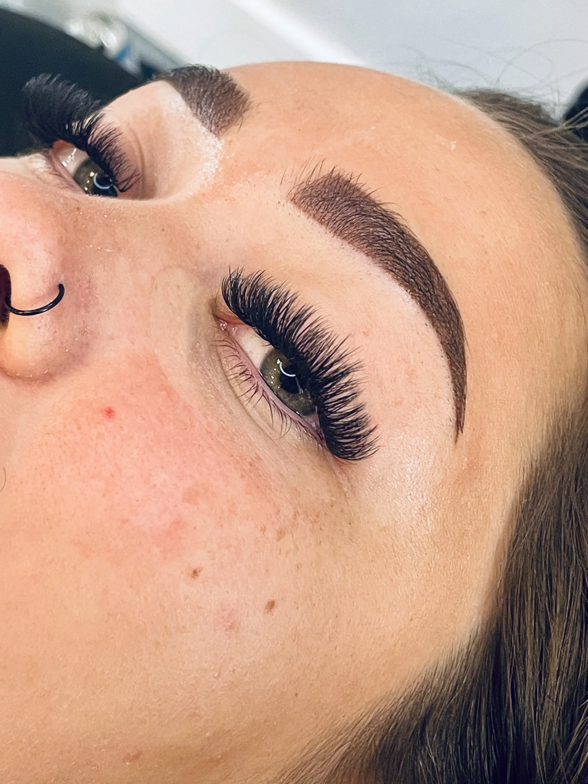 Close-up of a person's face showing eyebrows with microblading, long eyelashes, eye with makeup, and facial skin with minor blemishes.