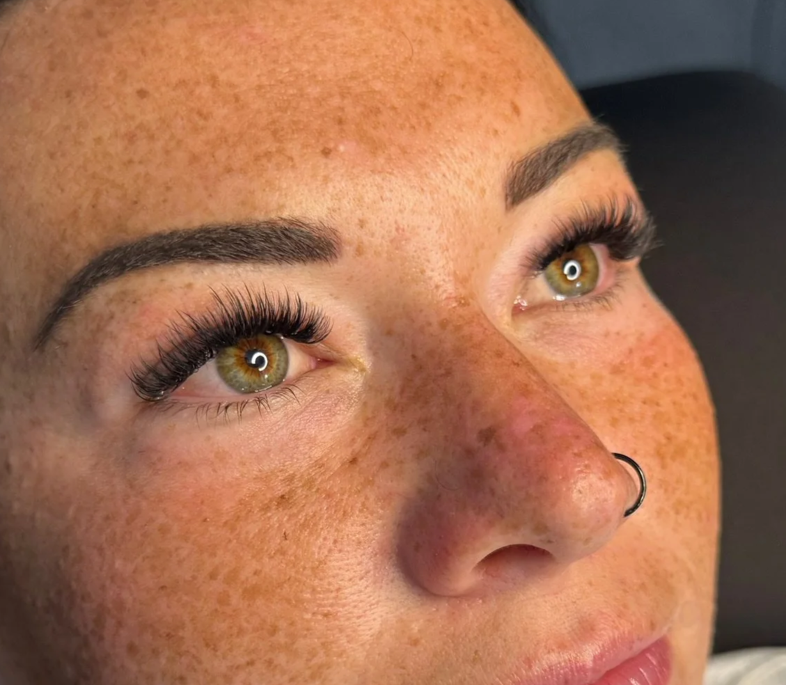 Close-up of a woman’s face showing her hazel eyes, dark long eyelashes, and a nose piercing. She has freckles and smooth skin.