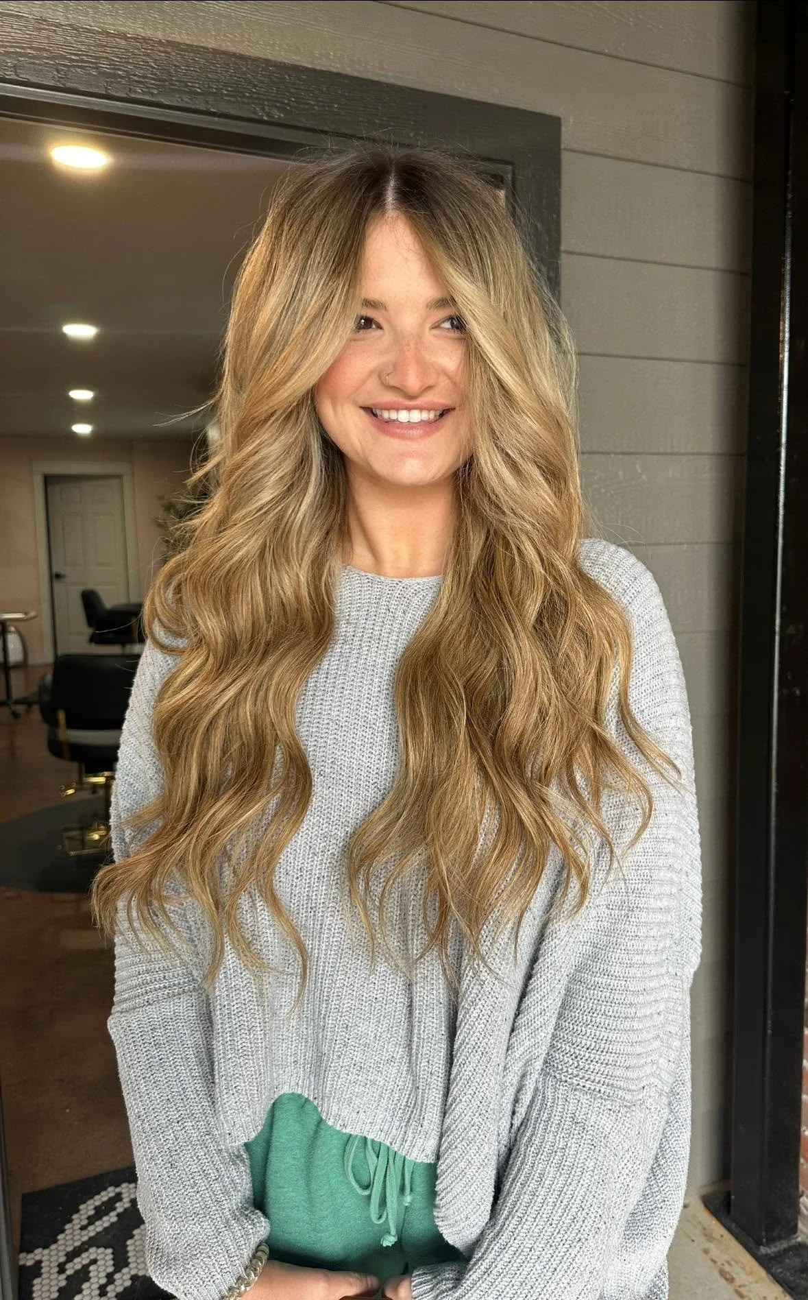 A woman with long wavy blonde hair, wearing a light gray sweater and green sweatpants, smiling indoors.
