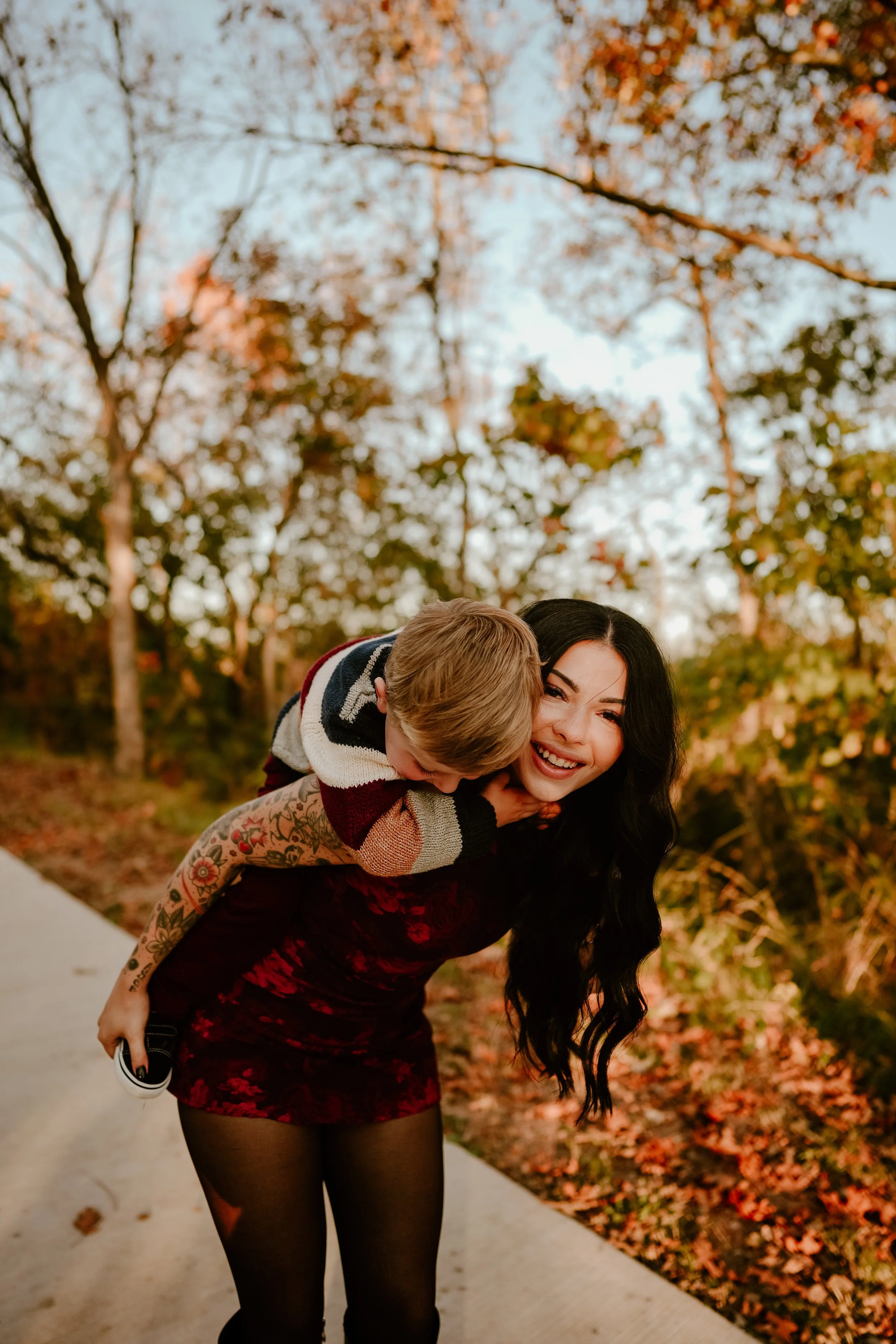 A woman with long black hair giving a piggyback ride to a young boy on an autumn day. They are outdoors on a pathway with fallen leaves, trees with orange and yellow leaves, and a clear sky in the background. The woman is smiling, wearing a red floral dress with black tights, and the boy is wearing a striped sweater and holding a shoe in his hand.