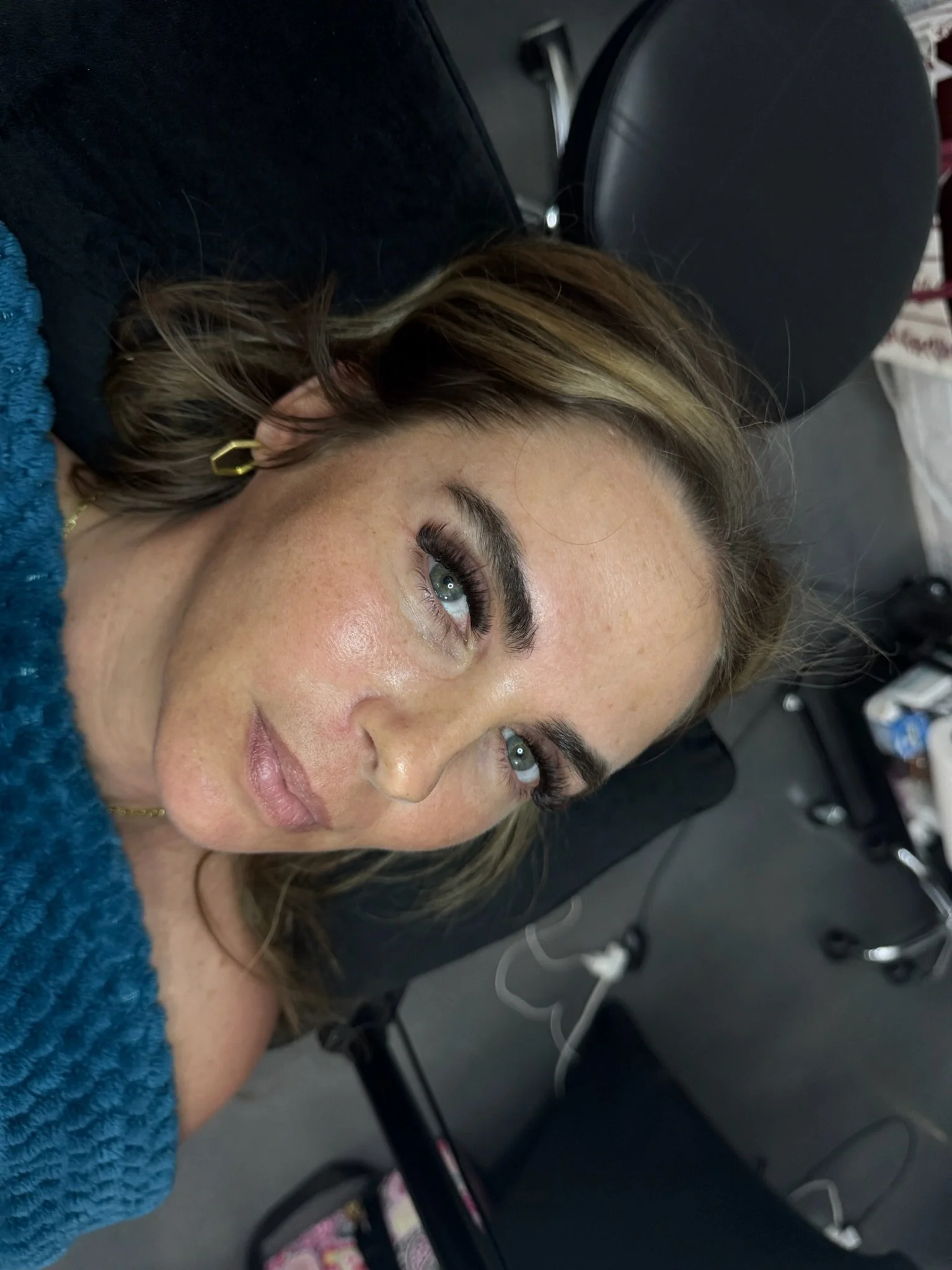 A woman with light skin, blue eyes, and brown hair with blonde highlights, lying on her side on a black chair, with a focus on her face and eye makeup, including painted eyelashes.