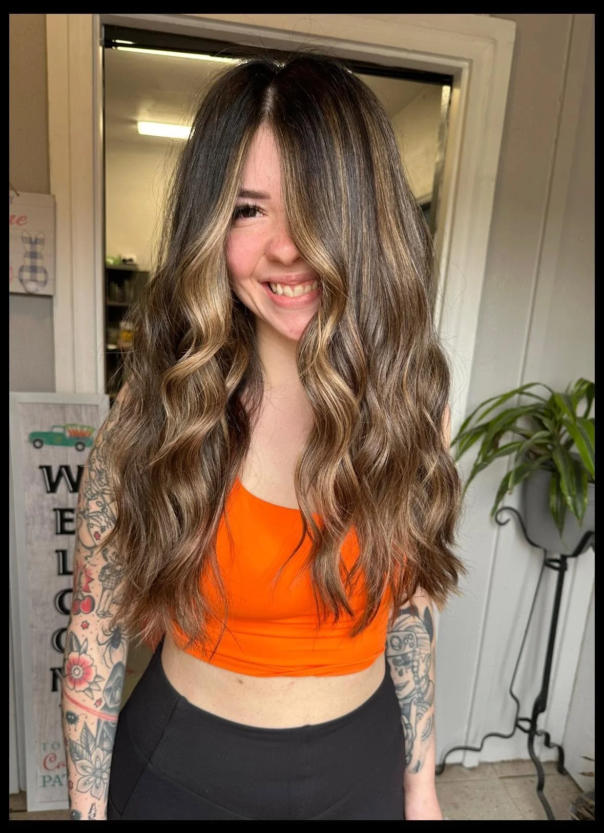 A woman with long, wavy hair dyed with blonde highlights and dark roots, smiling and wearing an orange crop top and black pants. She has tattoos on her arm and is standing indoors near a mirror and a plant.