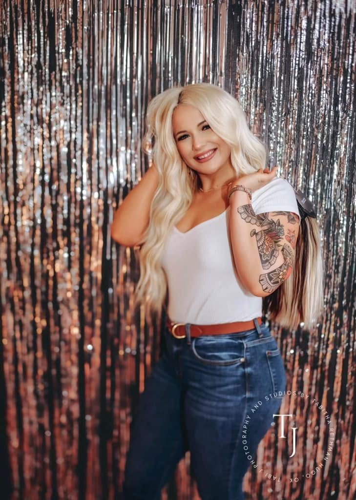 A smiling woman with long blonde hair and tattoos on her right arm, wearing a white t-shirt, blue jeans, and a brown belt, standing in front of a shimmering metallic backdrop.