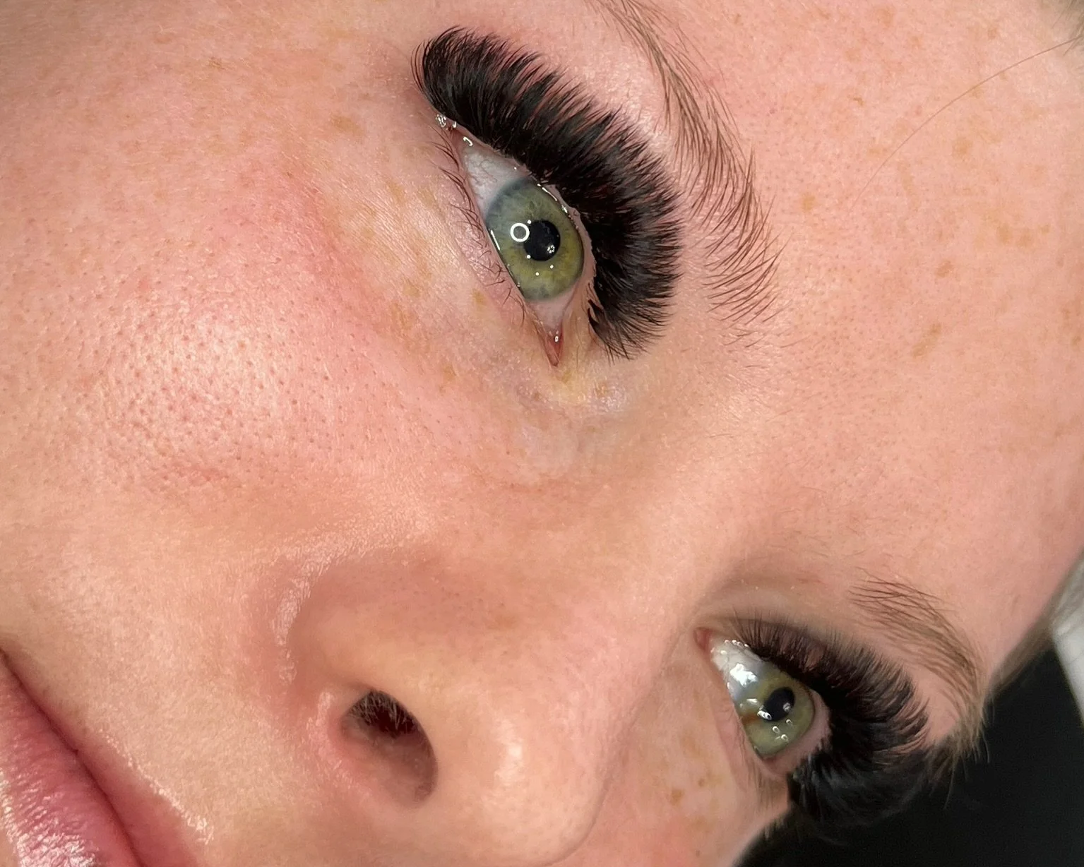 Close-up of a woman's face with green eyes, long black eyelashes, and light skin with a few freckles.