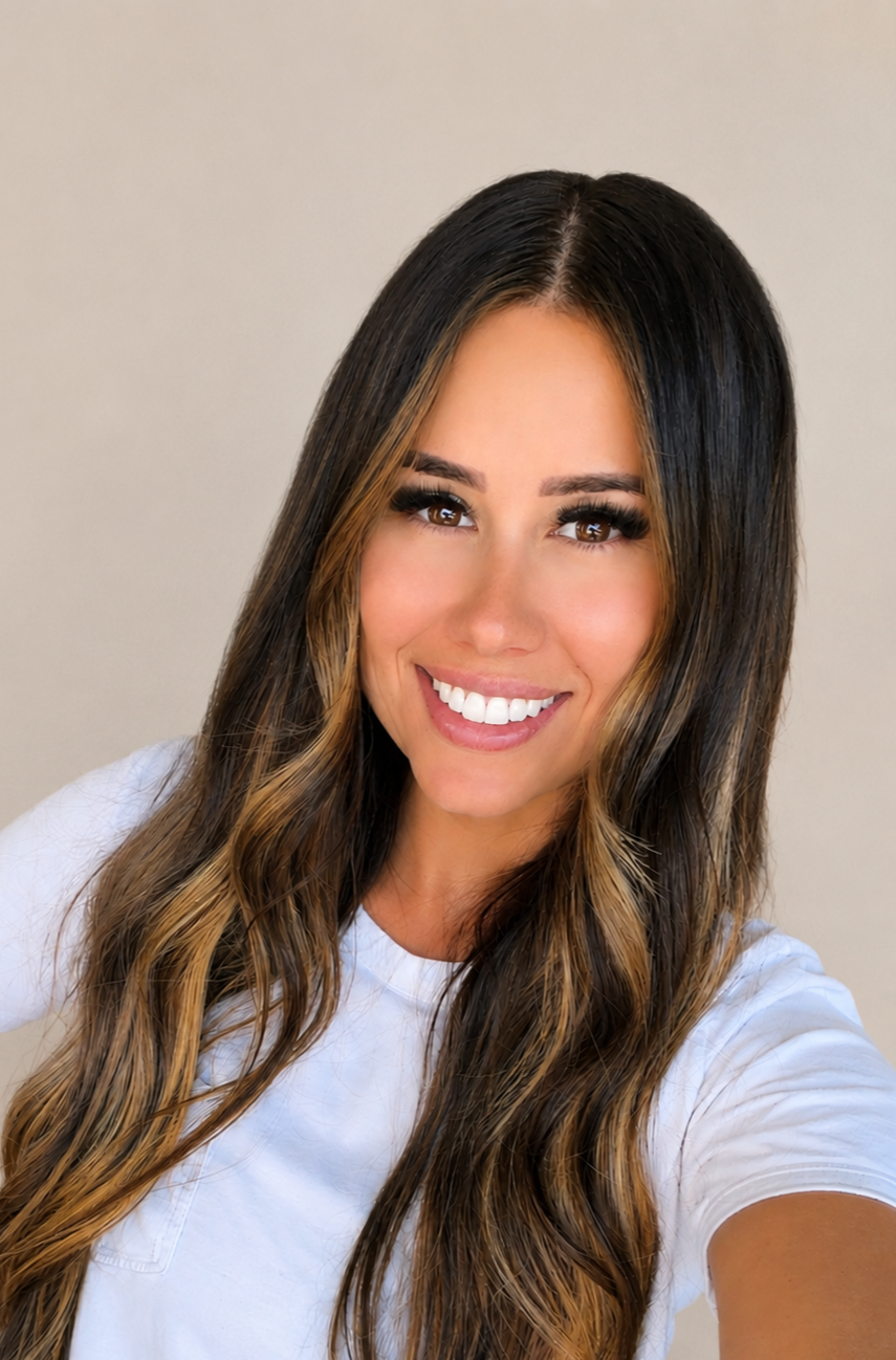 A woman with long wavy brown hair and light highlights, smiling at the camera, wearing a white shirt, against a neutral background.