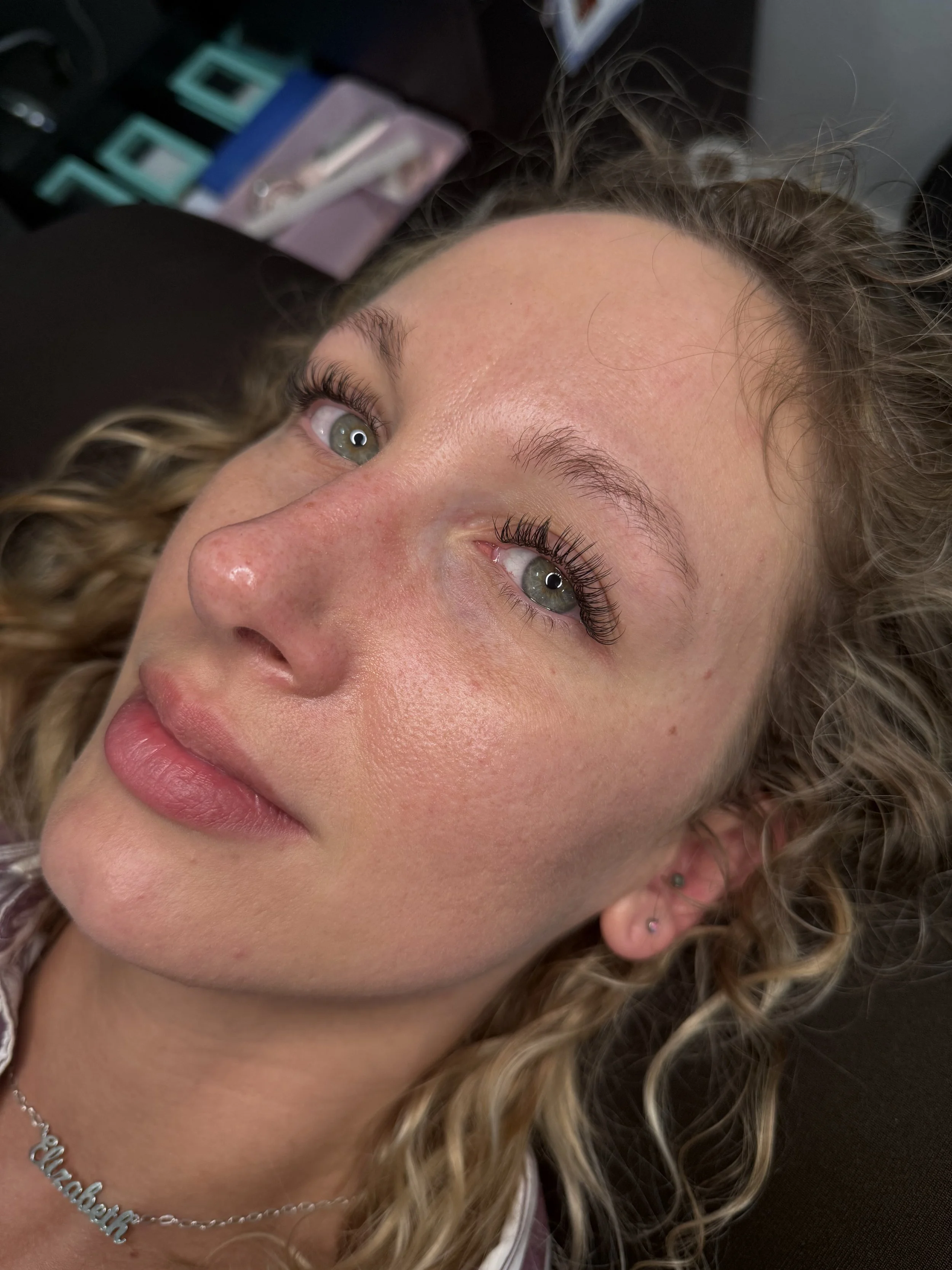 Close-up of a young woman's face lying down, showing her green eyes, curly blonde hair, and jewelry including earrings, a necklace with a nameplate, and a small nose piercing.