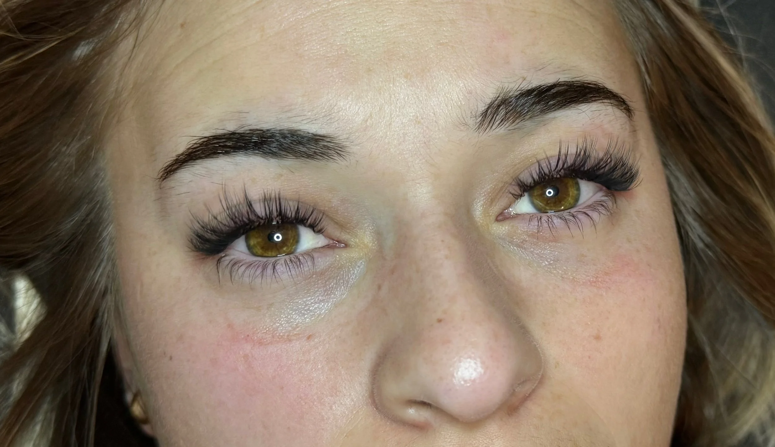 Close-up of a woman's face, showing her eyes, eyebrows, and part of her hair, with makeup including long eyelashes and well-groomed eyebrows.