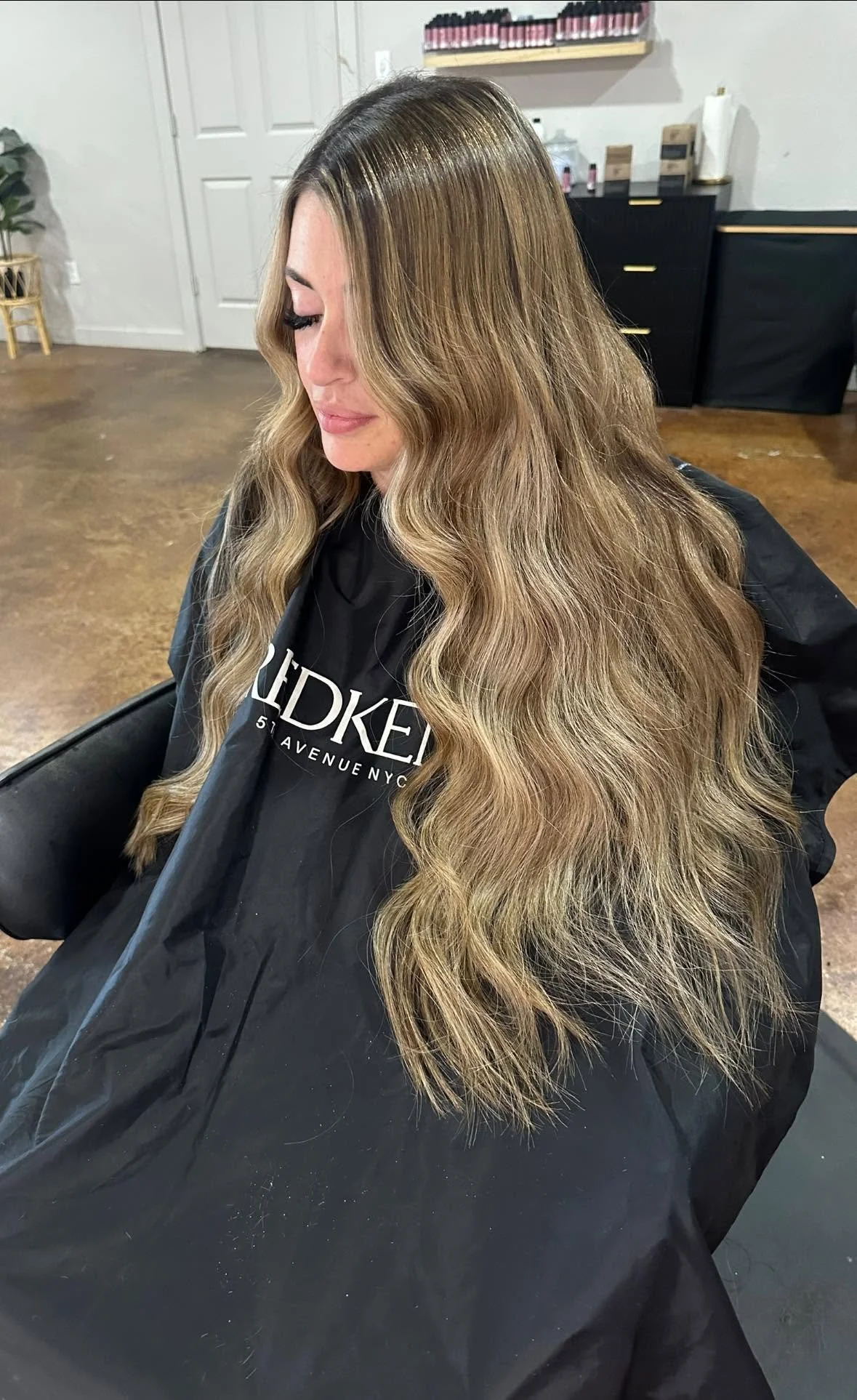 A woman with long wavy blonde hair sitting in a salon chair, wearing a black cape with white text. The salon has a hardwood floor, black furniture, and shelves with nail polish bottles.