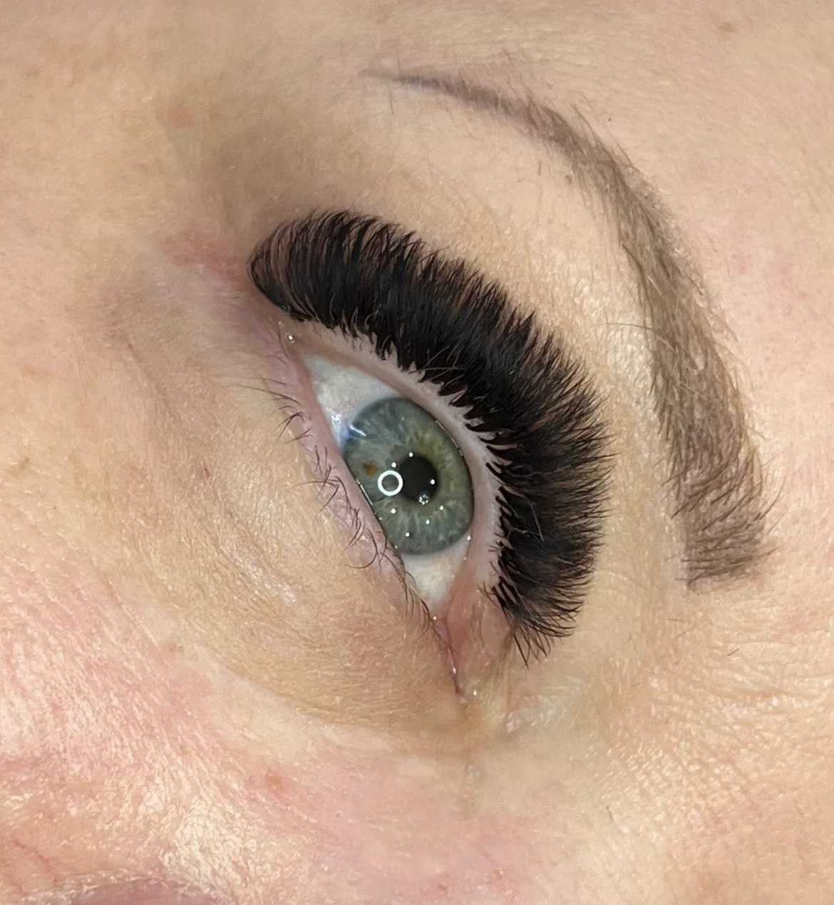 Close-up of a woman's eye with long, thick eyelash extensions, a green iris, and well-groomed eyebrow.