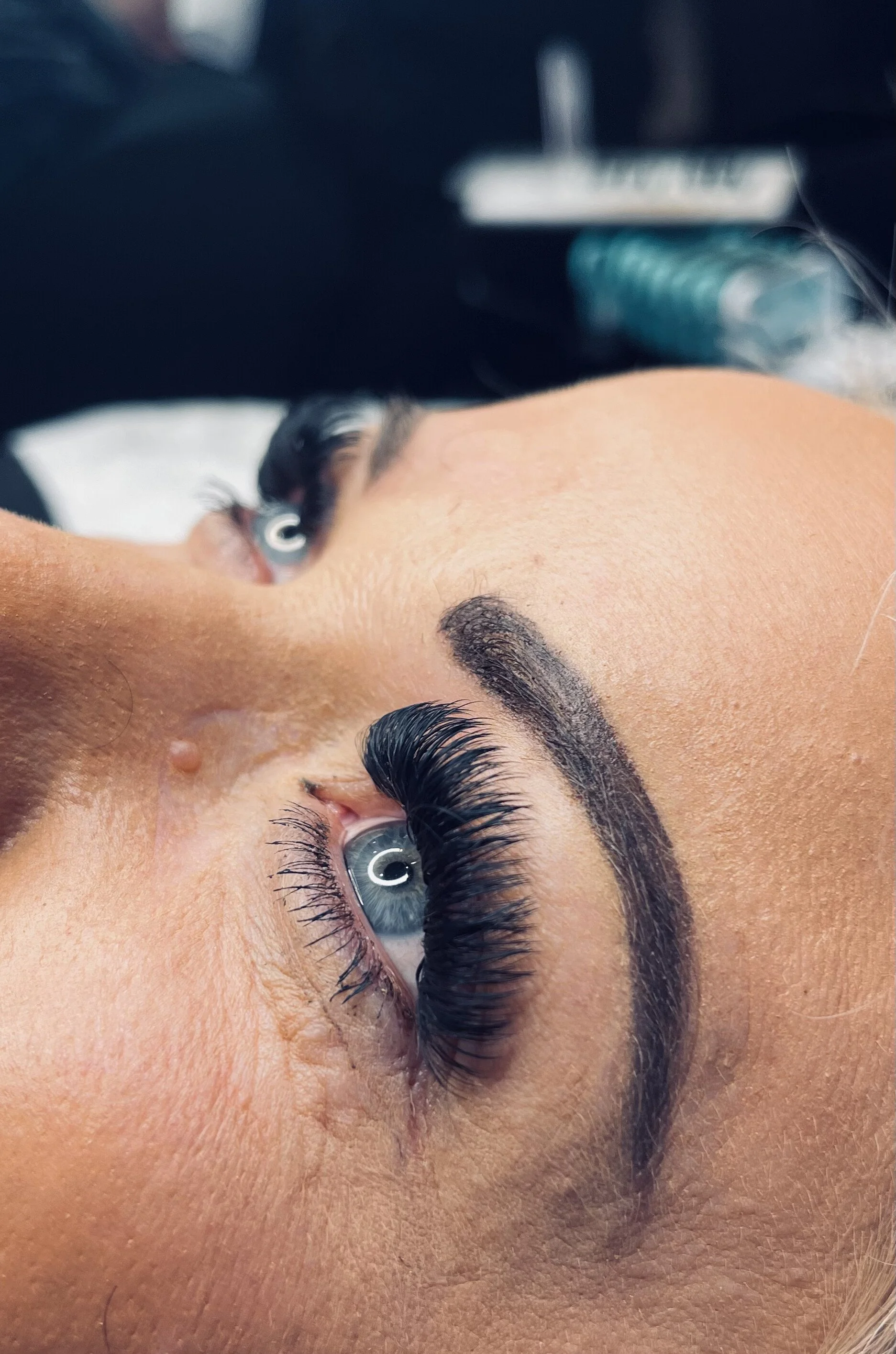 Close-up of a woman's face showing her eye with long, curled eyelashes, dark eyebrows, and a natural look. The woman appears to be receiving a beauty treatment.
