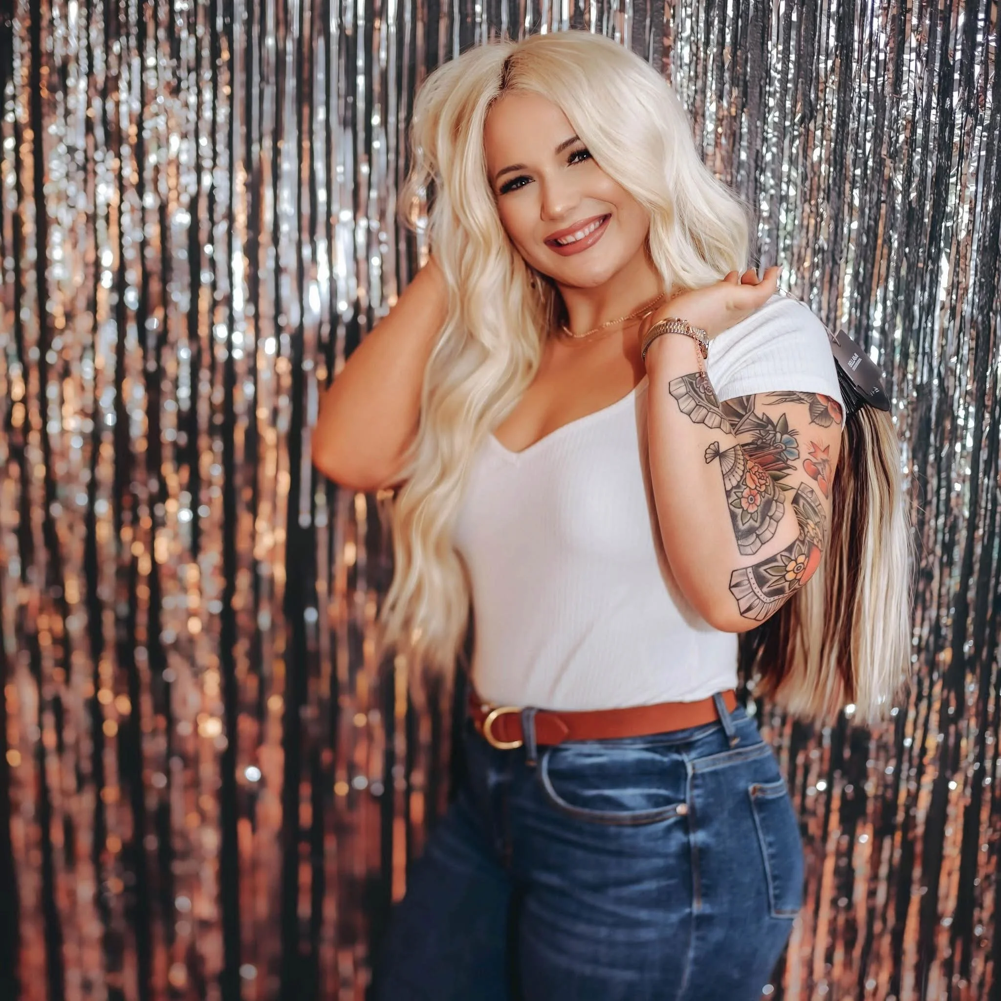 A woman with long blonde hair, smiling, wearing a white shirt and blue jeans, standing in front of a shiny silver curtain backdrop, showing a floral tattoo on her upper arm and wrist jewelry.