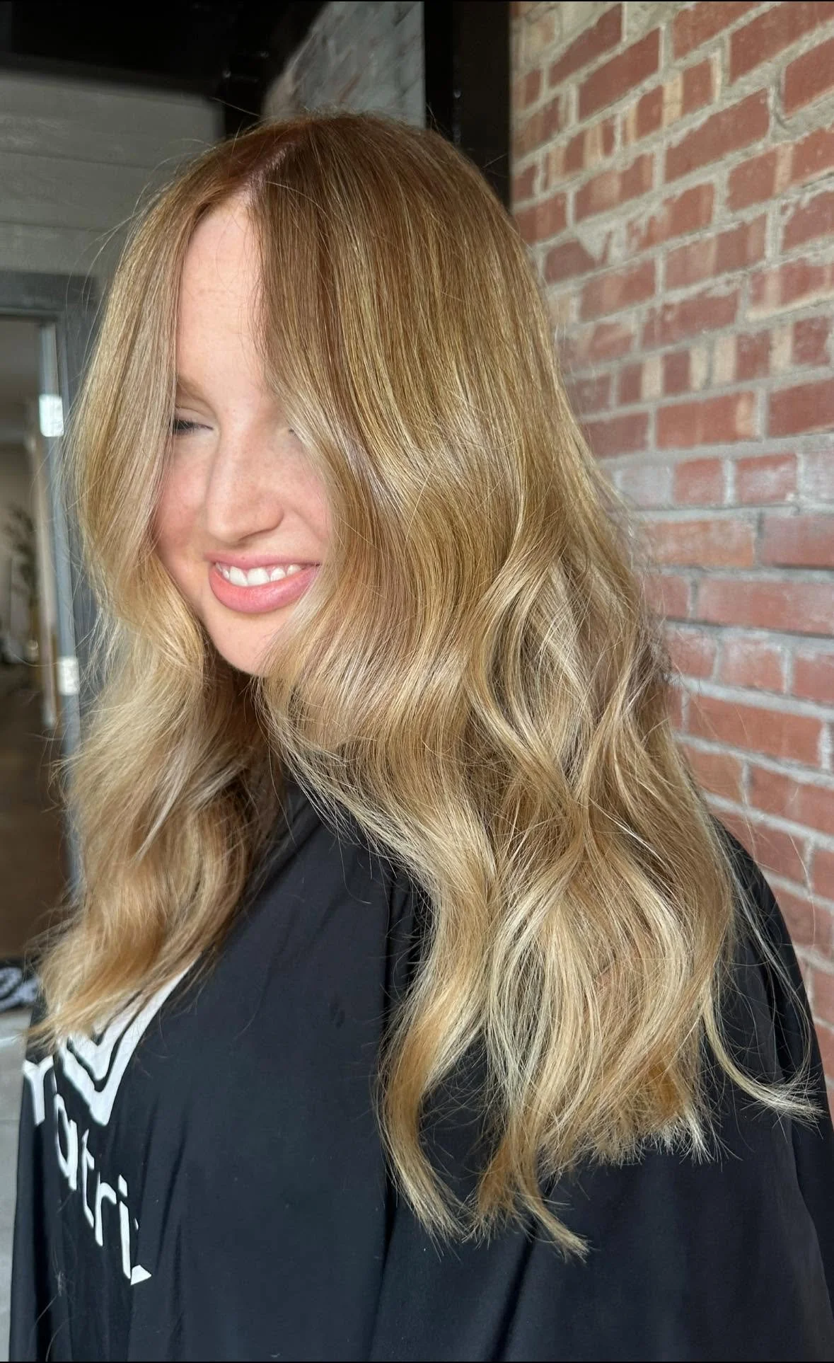 Close-up of a woman with long, wavy blonde hair standing indoors in front of a brick wall, smiling with eyes closed.