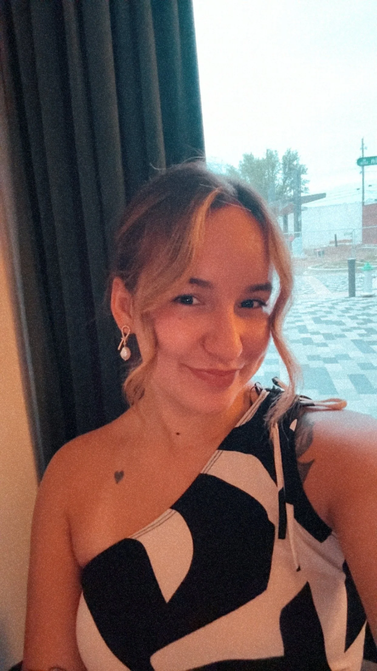 A woman with wavy, blonde hair and earrings, smiling for a selfie inside a building near a window with a view of an outdoor area and street signs.