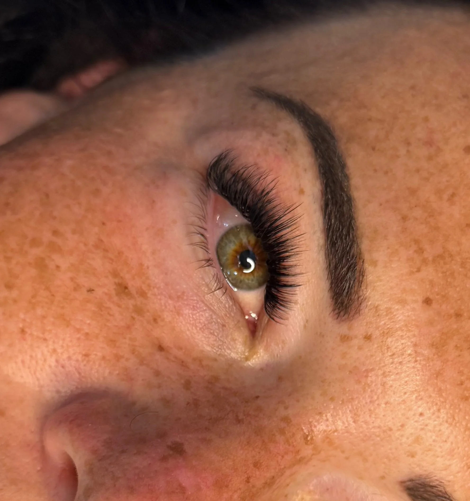 Close-up of a person's eye, showing hazel iris, dark eyelashes, and brown eyebrow, with freckles and skin texture surrounding the eye.