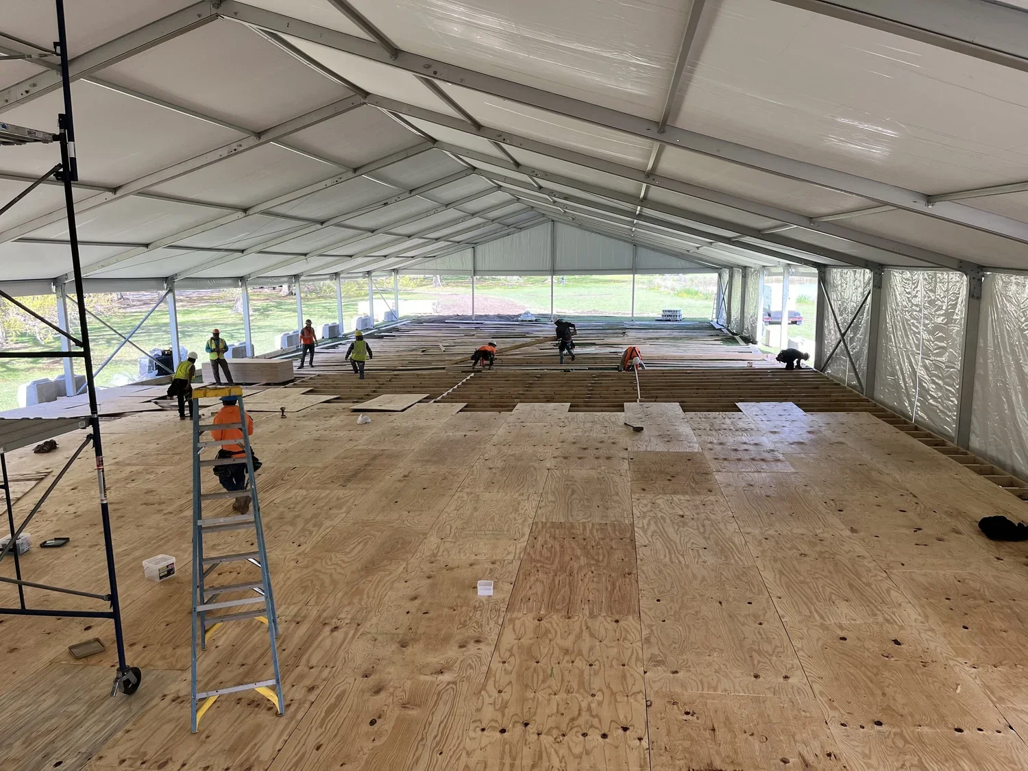 Workers installing wooden flooring inside a large job site tent with open sides, supported by a metal framework, during the daytime for Larkin's Industrial Tents.
