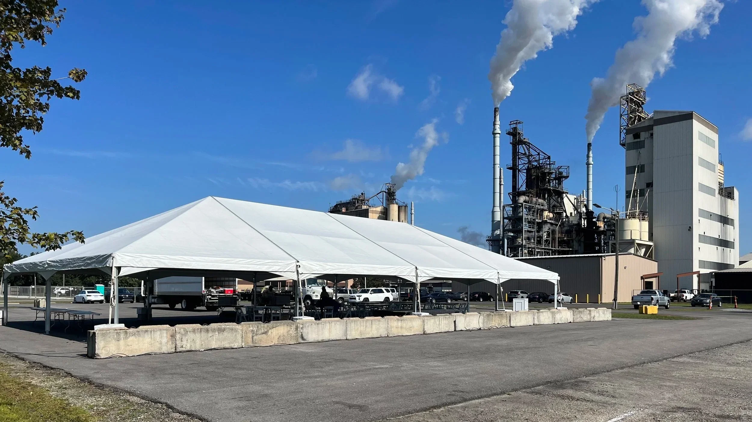 A large industrial facility with smokestacks emitting smoke or steam, partly obscured by a white event tent set up in a parking lot, with parked cars and trees around.