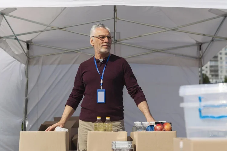 A man with gray hair and glasses stands behind a table with bottled water, apples, and other supplies under a white canopy tent for Larkin's Industrial Tents.