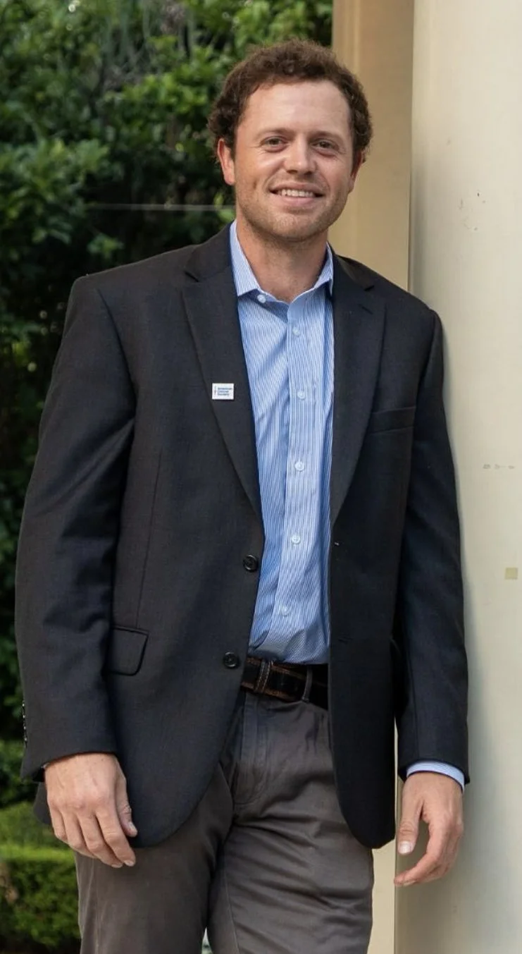 A man with curly hair wearing a dark gray suit jacket, light blue dress shirt, and dark pants, standing outdoors next to a beige column, with green foliage in the background.