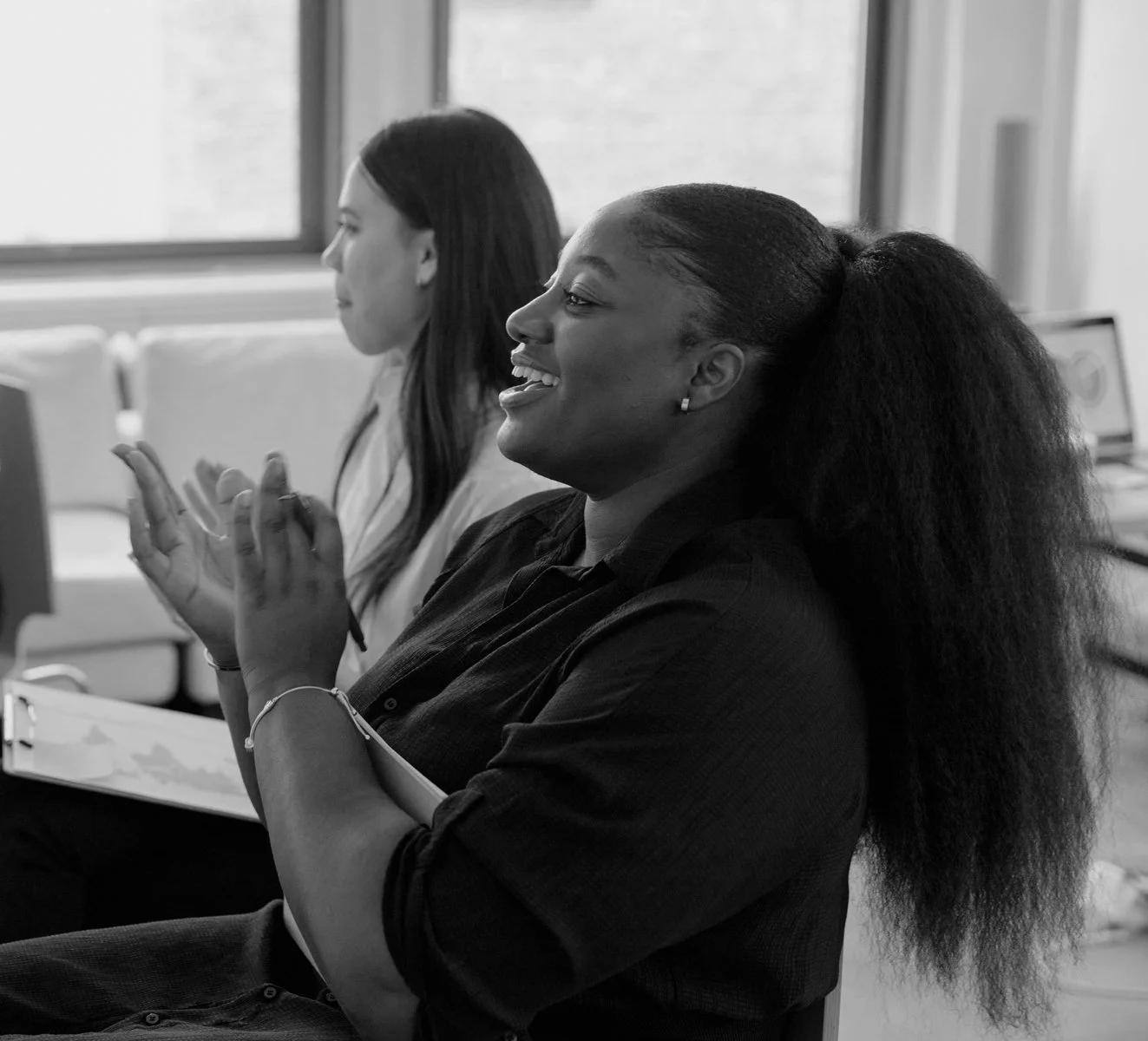 Two women sitting in a classroom or meeting room, clapping and smiling, one with long natural hair, the other with straight hair, near a window.