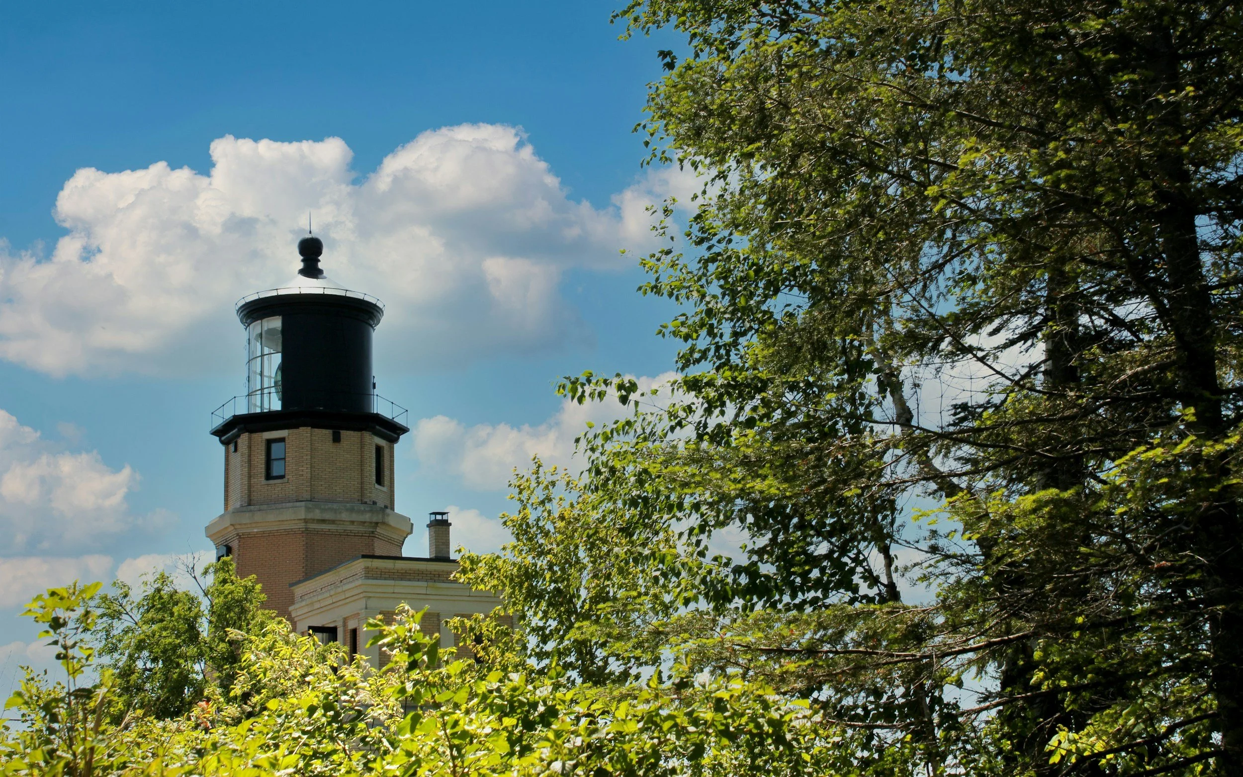 A lighthouse with a black top and beige brick base, partially obscured by green tree branches and leaves, under a blue sky with white clouds.