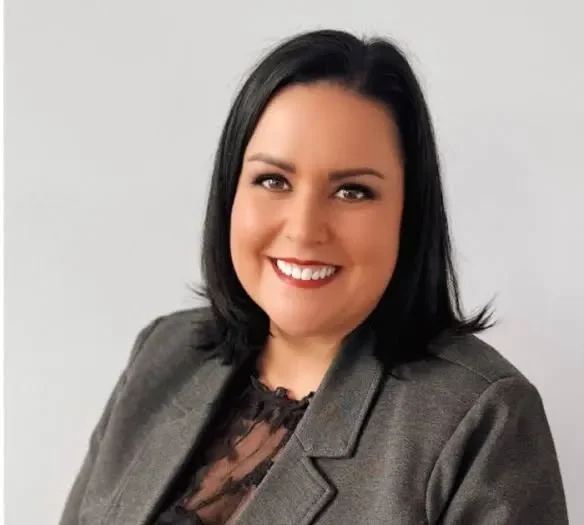 A woman with shoulder-length black hair smiling, wearing a gray blazer and black lace top, against a plain light gray background.
