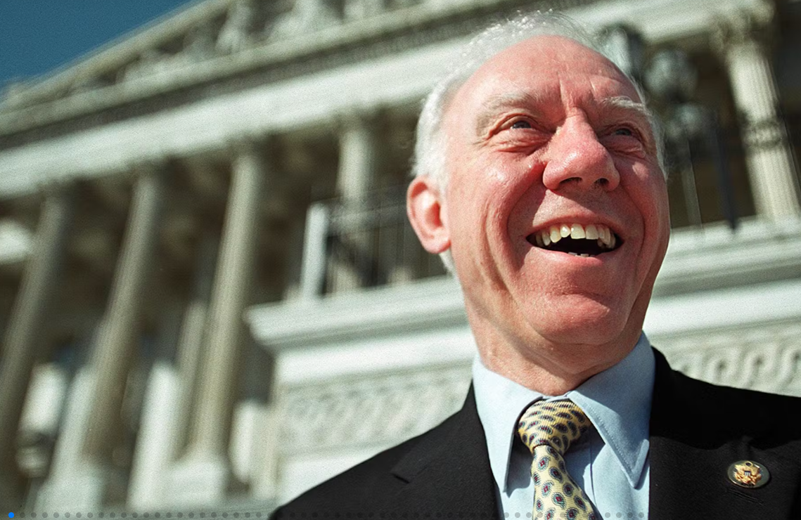 Smiling elderly man in a suit with a patterned tie standing in front of a large, historic building with columns.